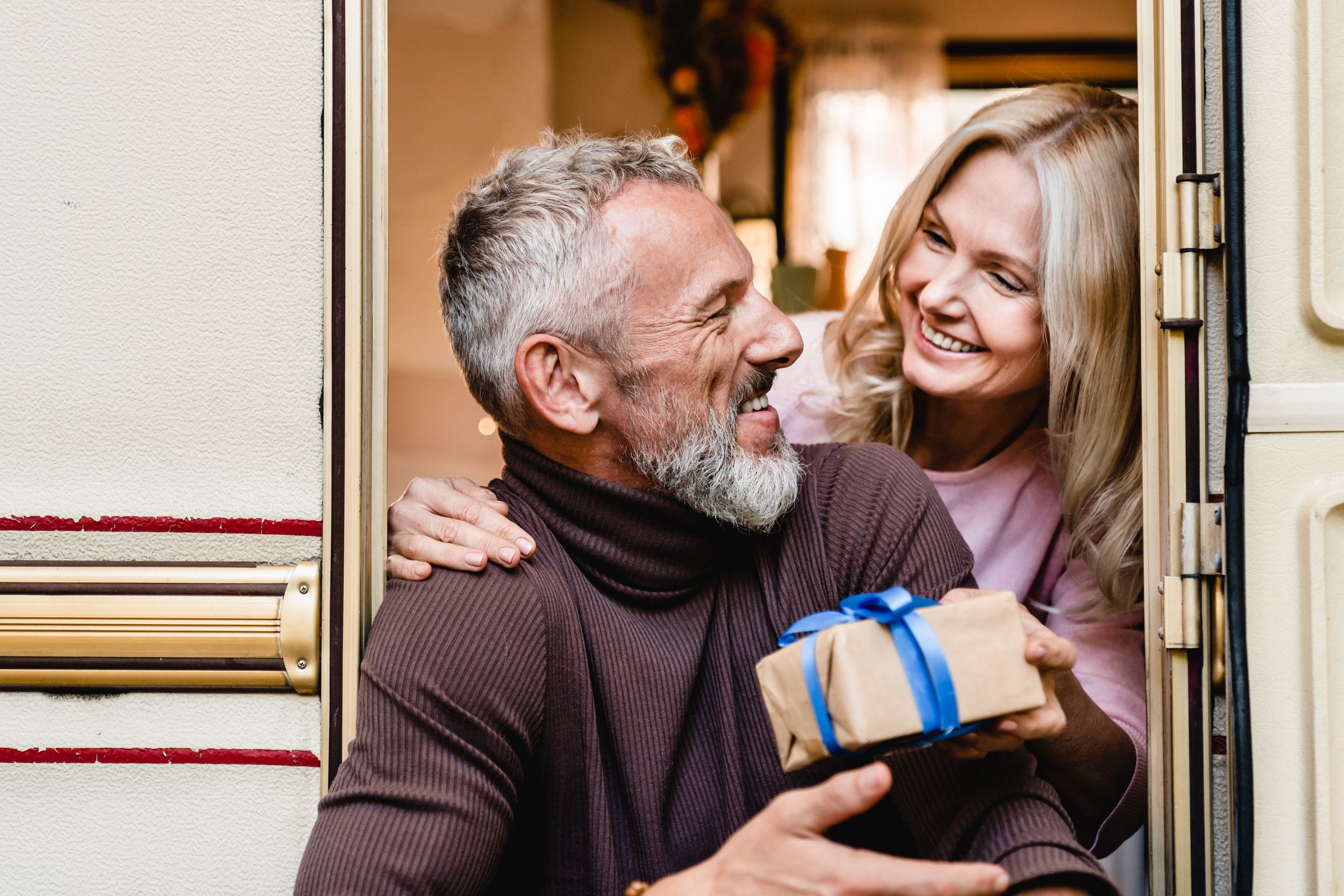 Blonde senior beautiful woman giving her grey-haired husband a present in their mobile home