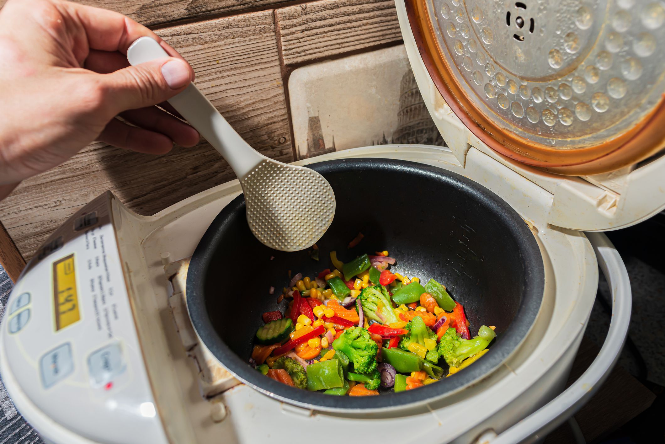 mixed set of different vegetarian vegetables in a white multicooker is stirred with a kitchen plastic Scraper. Onions, peppers, paprika, peas, corn, broccoli, cabbage and cucumber
