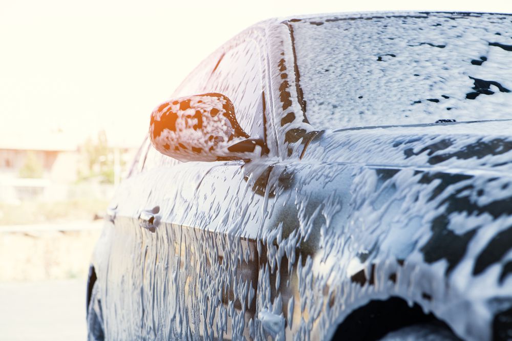 A close-up of a black car covered in white soap suds during a car wash, with sunlight illuminating the scene. The side mirror and windows are visible, partially obscured by the foam.
