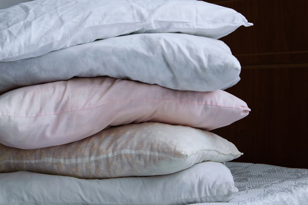 A stack of five pillows in different shades of white and light pink are piled on top of each other, resting on a white quilted mattress. The background includes a dark wooden surface.