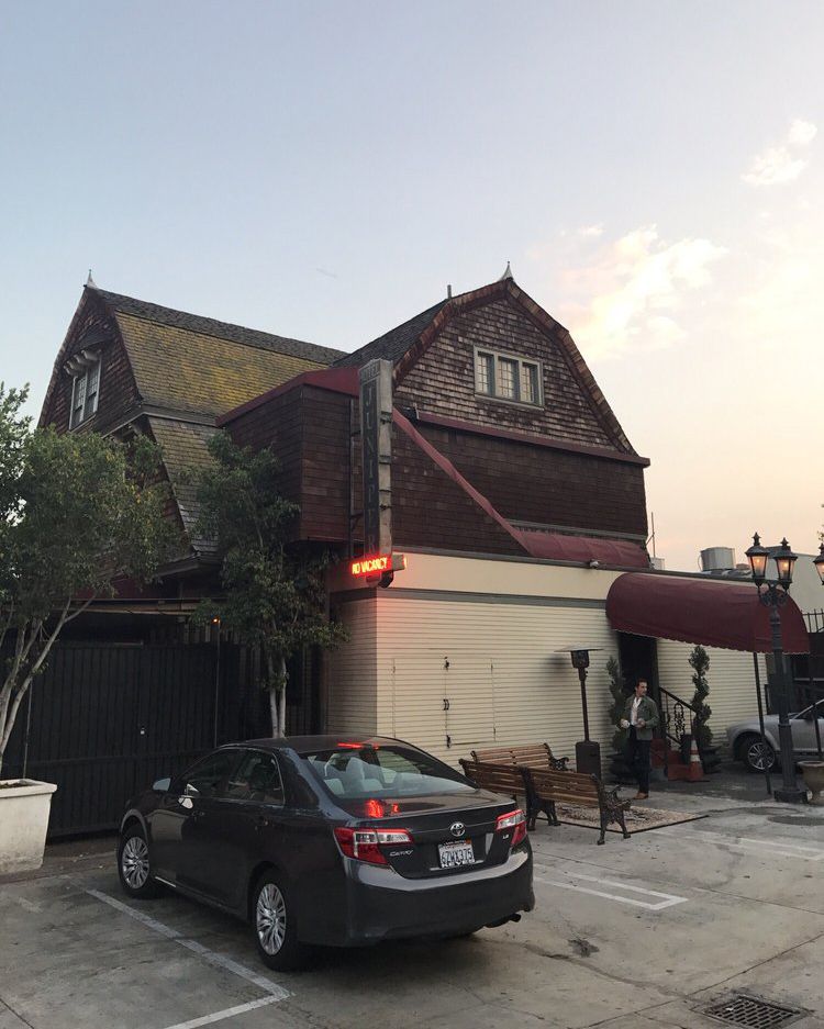 A dark sedan is parked in front of a rustic, brown two-story building with a red canopy and neon sign reading "Cocktail." Trees and benches line the sidewalk under a clear evening sky.
