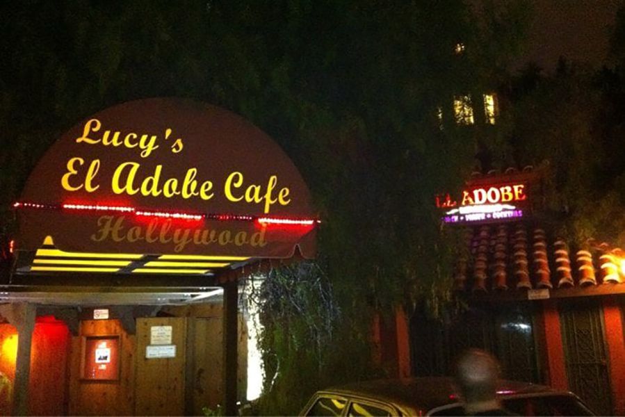 A nighttime view of Lucy’s El Adobe Cafe in Hollywood, showing illuminated yellow and red neon signs above the entrance and on the building, with a car parked in front and trees partially obscuring the scene.