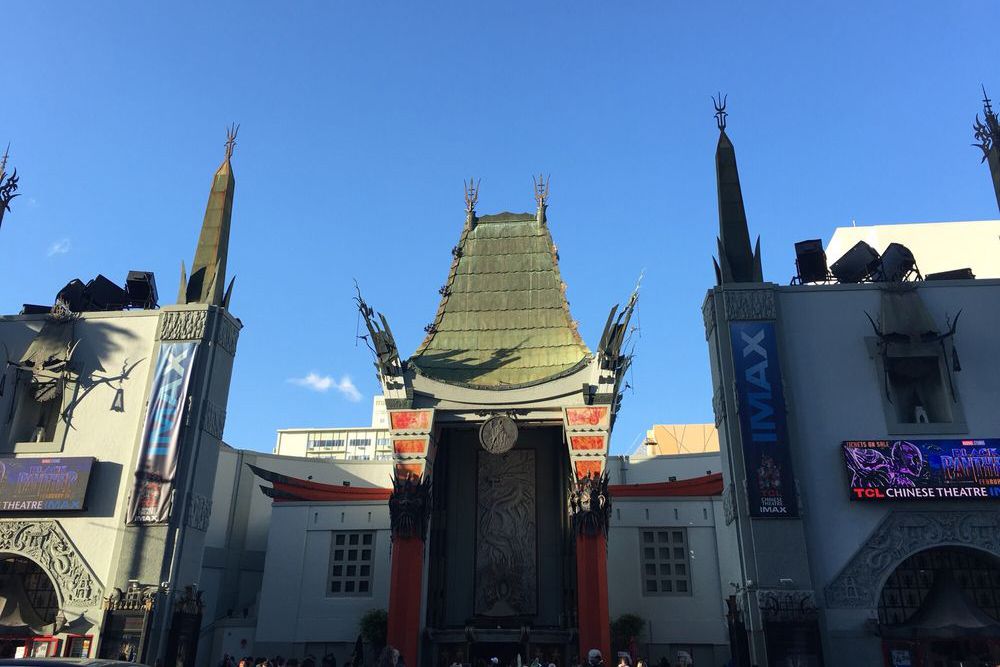 The exterior of TCL Chinese Theatre in Hollywood, featuring its ornate roof and decorative towers, with clear blue sky above and digital movie billboards on both sides of the entrance.