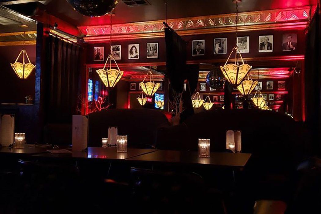 Dimly lit lounge with ornate hanging lamps, red accent lighting, framed black-and-white photos on the walls, candles on tables, and mirrors reflecting the cozy, elegant atmosphere.