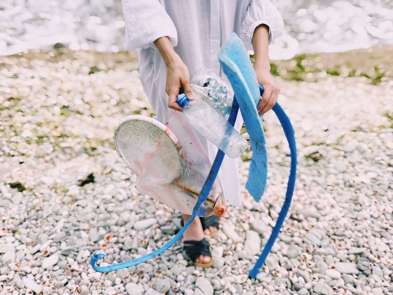 A person standing on a rocky shore holds various pieces of plastic litter, including bottles and blue debris, likely collecting trash to clean up the environment.