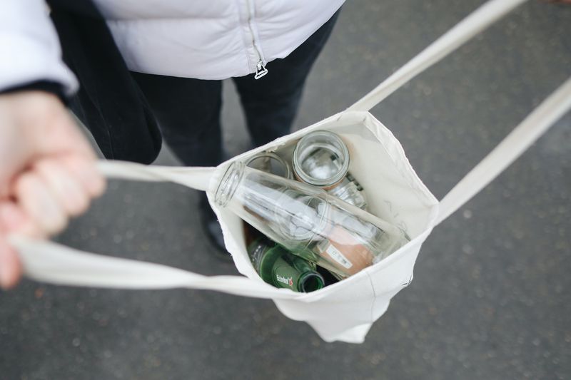 A person holds a white tote bag filled with empty glass bottles and jars, viewed from above, standing on a gray pavement.