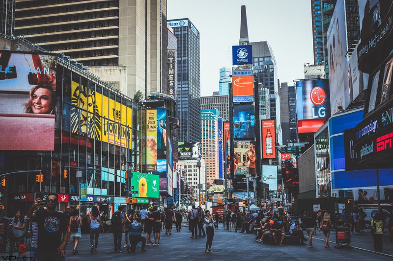Crowds of people walk through Times Square in New York City, surrounded by tall buildings and colorful digital billboards displaying advertisements and bright lights.