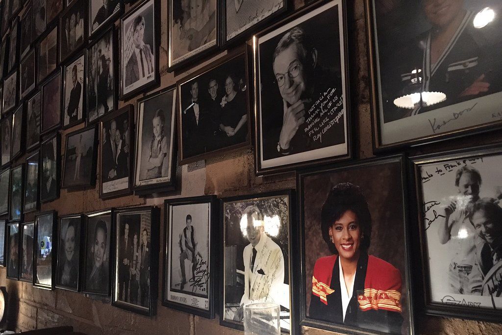 Framed black-and-white and color photographs of various people, some autographed, cover a wall in a dimly lit room, creating a collage-like display.
