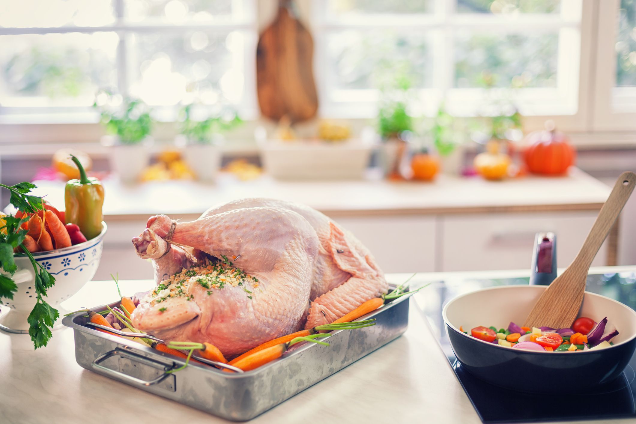 A raw turkey in a metal baking pan, surrounded by carrots and other vegetables, on an island in the kitchen, next to a skillet with cut vegetables and a wooden spatula, on the stove, with the kitchen countertop and a large window with sunlight coming thro