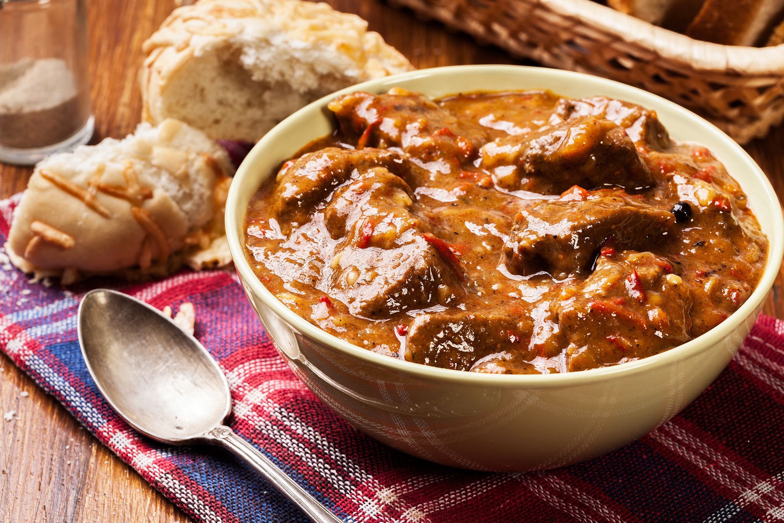 All-American beef stew in a white ceramic bowl next to a rustic spoon on a red and blue plaid napkin with two pieces of bread, on a wooden table, blurred background