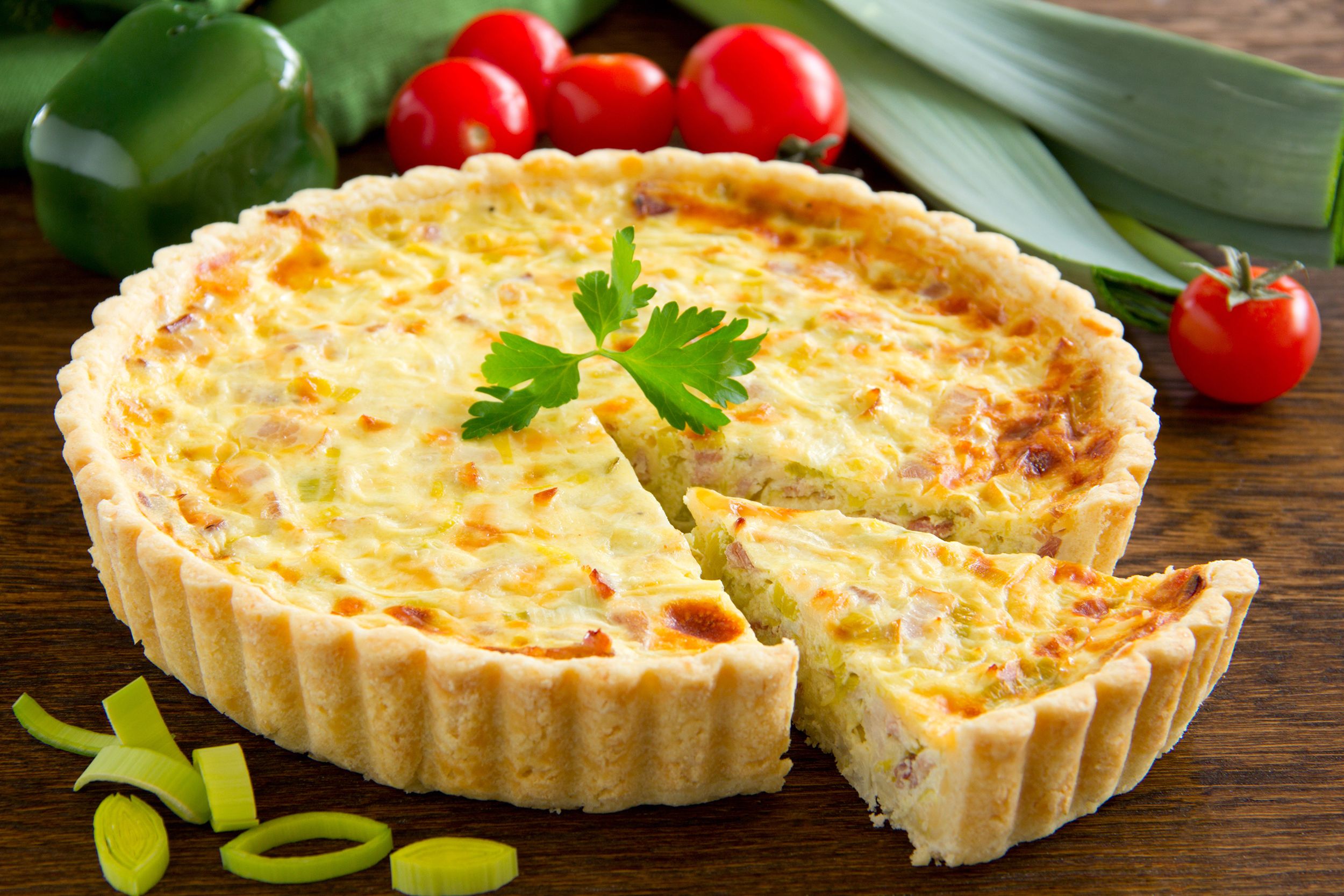 Asparagus, spinach, & feta quiche with one piece cut and separated, surrounded by ingredients such as cherry tomatoes and a green bell pepper on a wooden table