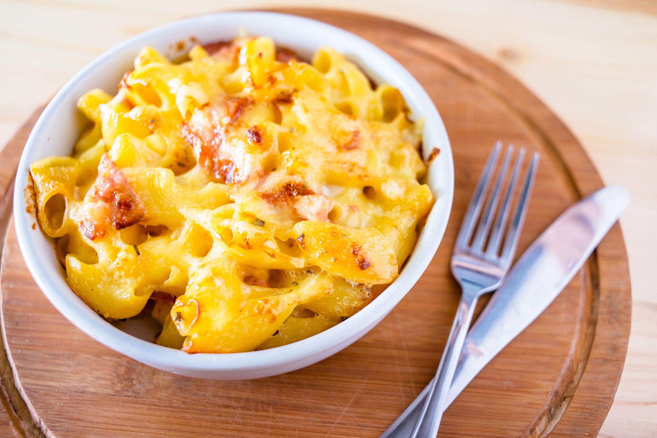 Bacon macaroni and cheese in a white bowl on the left, selective focus, with a fork and a knife on the right, slightly blurred on a round wooden plate, blurred background of a light wooden table