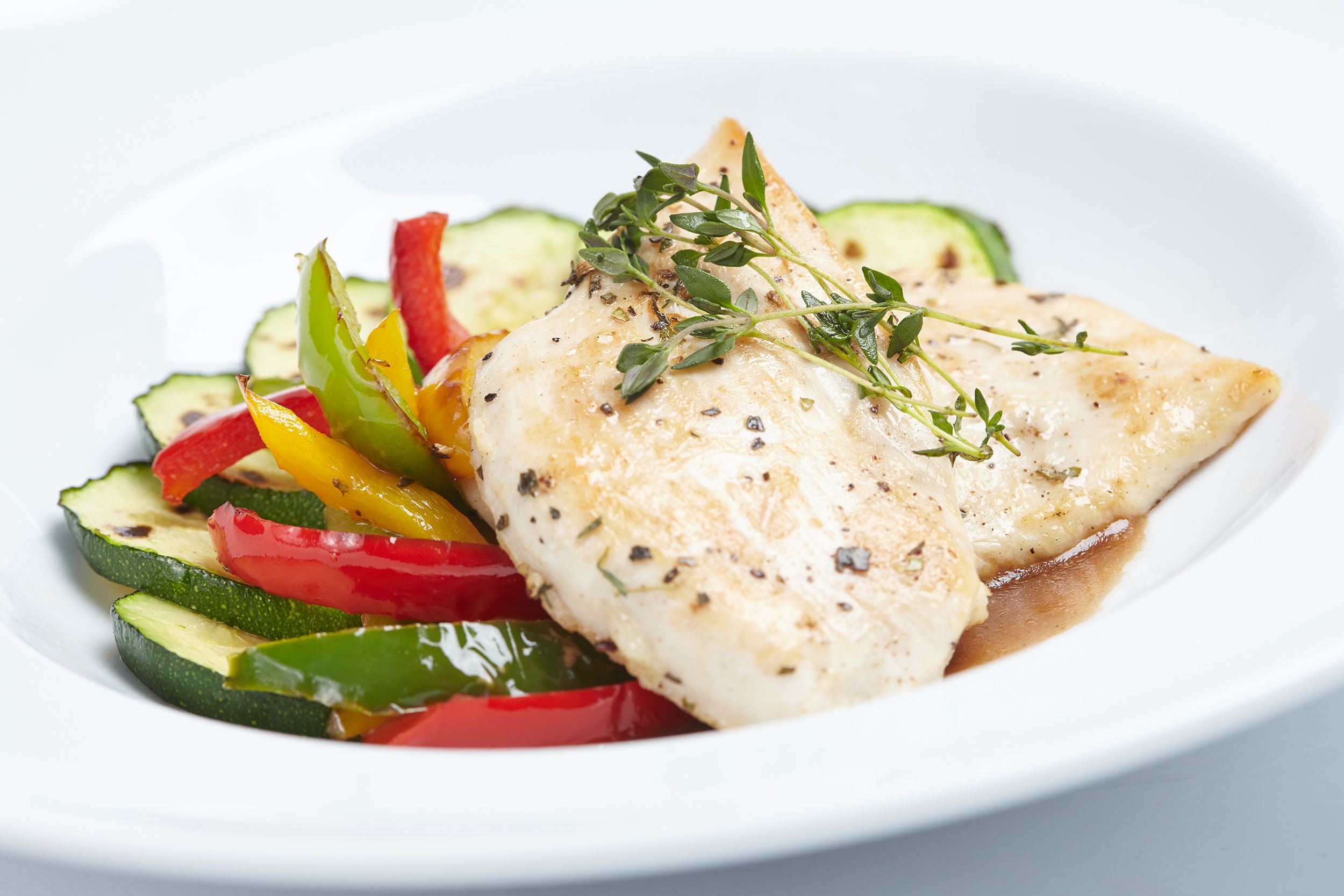 Baked bluefish with zucchini and sliced bell pepper in a white porcelain bowl, slight diagonal with a white background