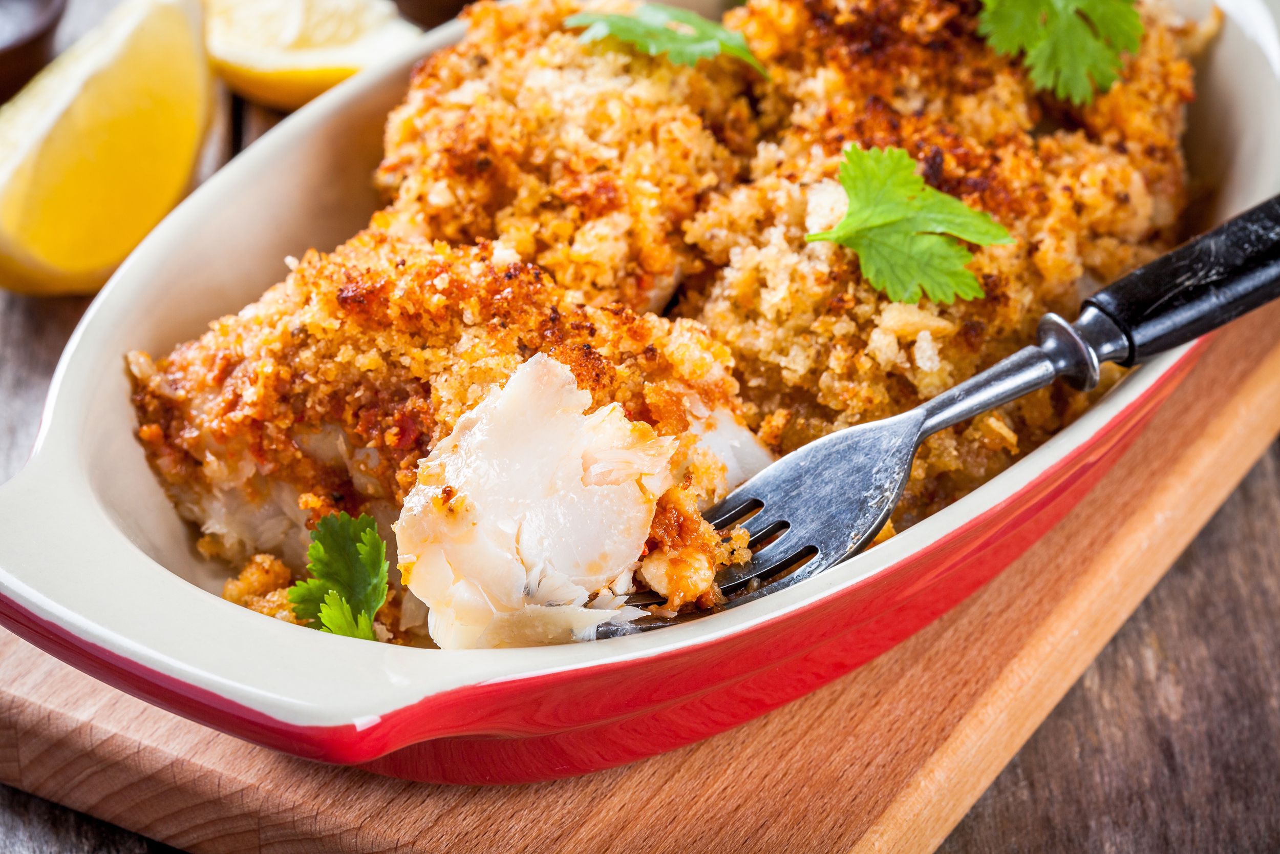 Baked cod with Ritz cracker topping in a red ceramic gratin dish with a fork holding a chunk of it, selective focus, on a wooden cutting board, surrounded by lemons
