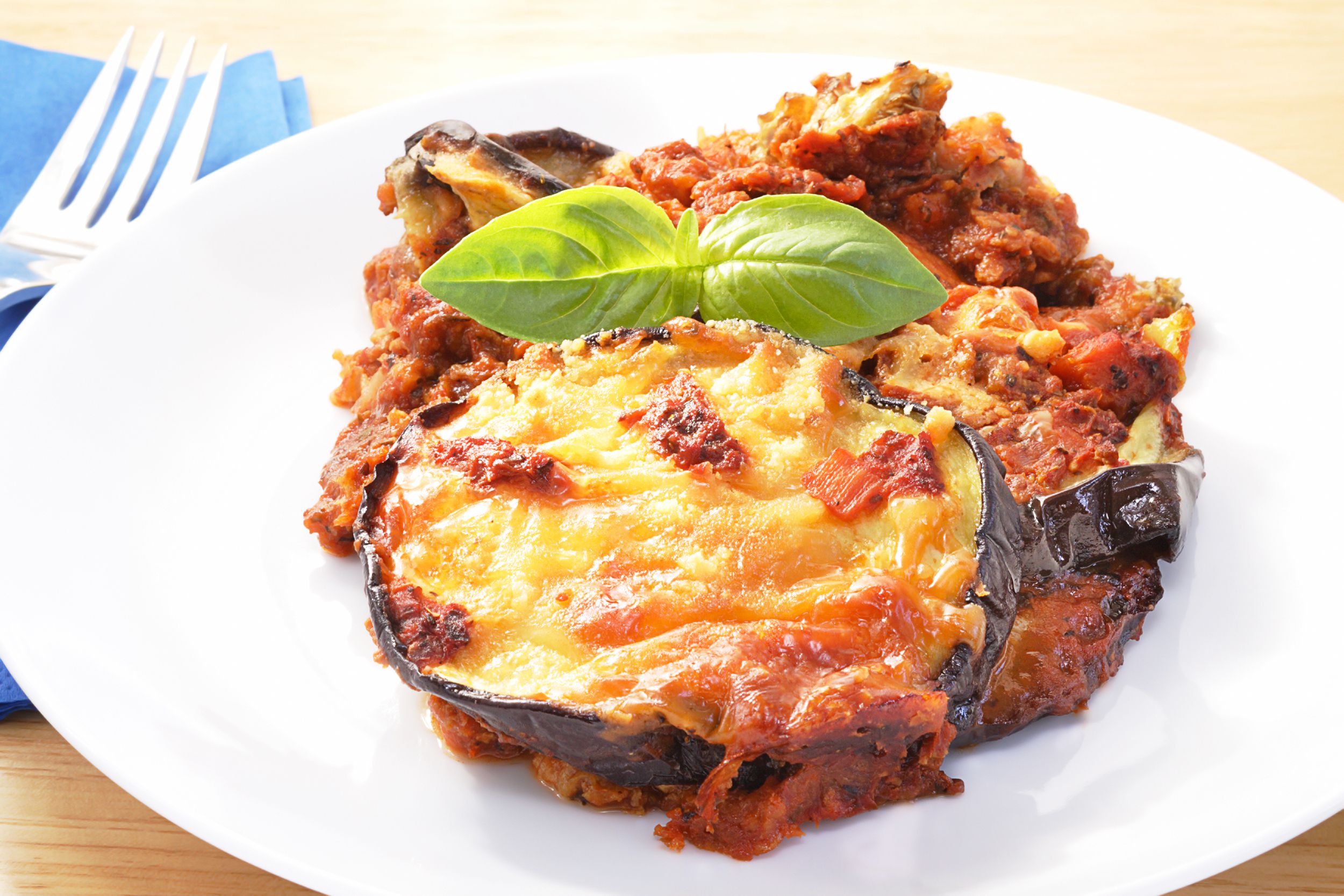 Baked-eggplant parmesan on a porcelain white plate with a fork on a blue napkin to the left on a table