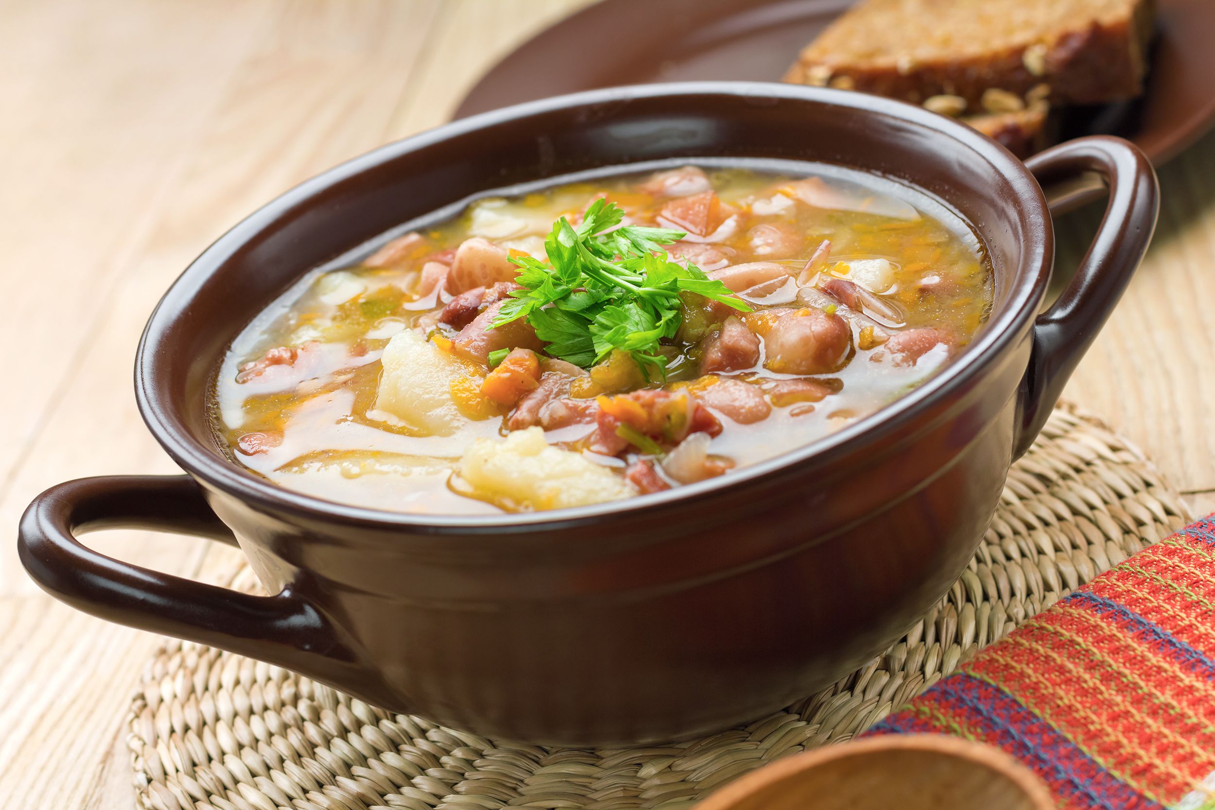 Bean with bacon soup in a brown ceramic bowl with two handles on each side, slight diagonal, on a round fiber table mat, with a matching brown ceramic plate with bread, all on a light wooden table