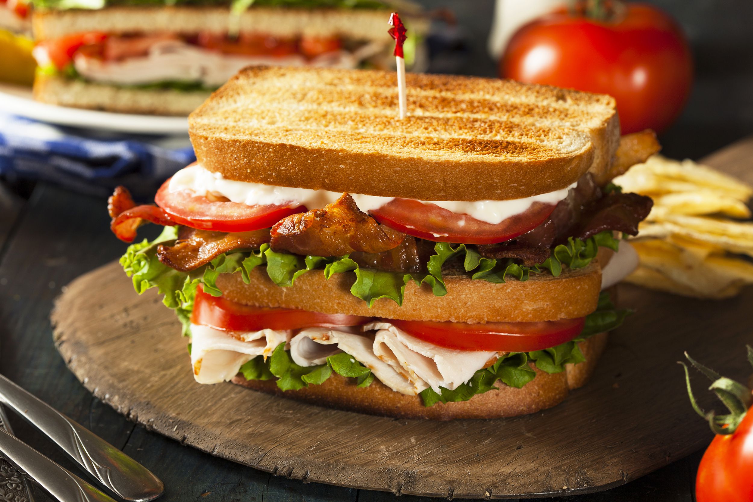 Classic turkey club on an artisanal round wooden serving board, selective focus, with another club sandwich and ingredients blurred in the background