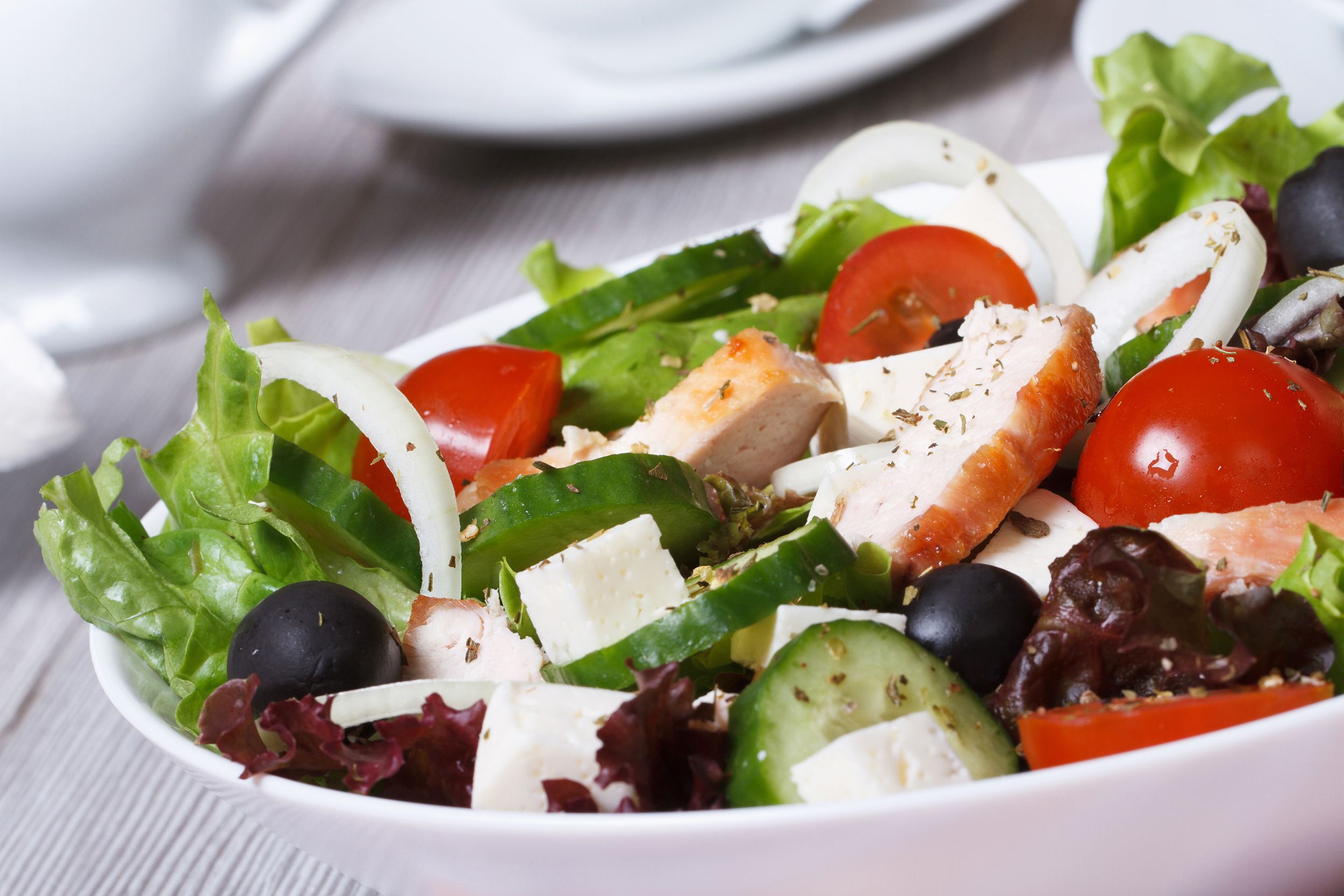 Closeup of chopped Greek salad with chicken in a white bowl on a grey wooden table with a blurred background of dishes