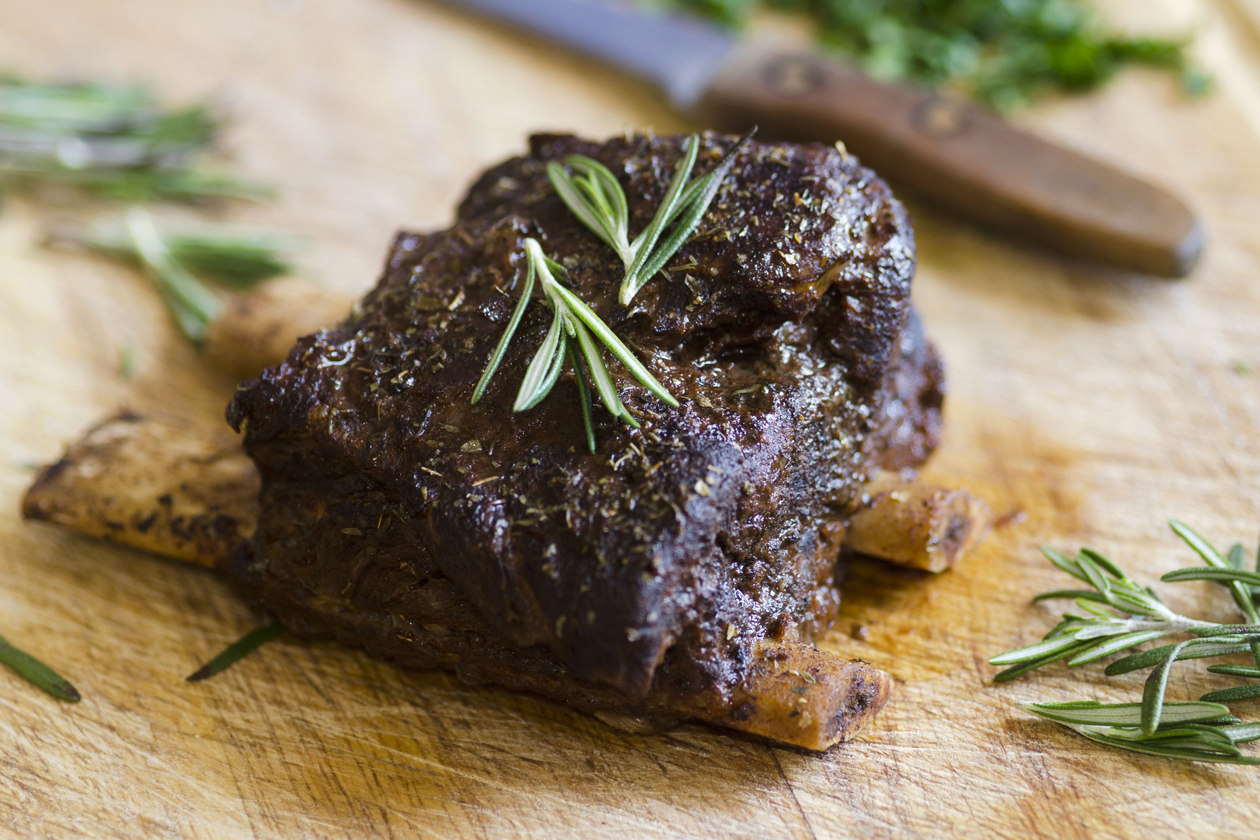 Closeup of easy BBQ short ribs on a wooden cutting board, with knife lines in view, with a knife and herbs blurred in the background, on the cutting board