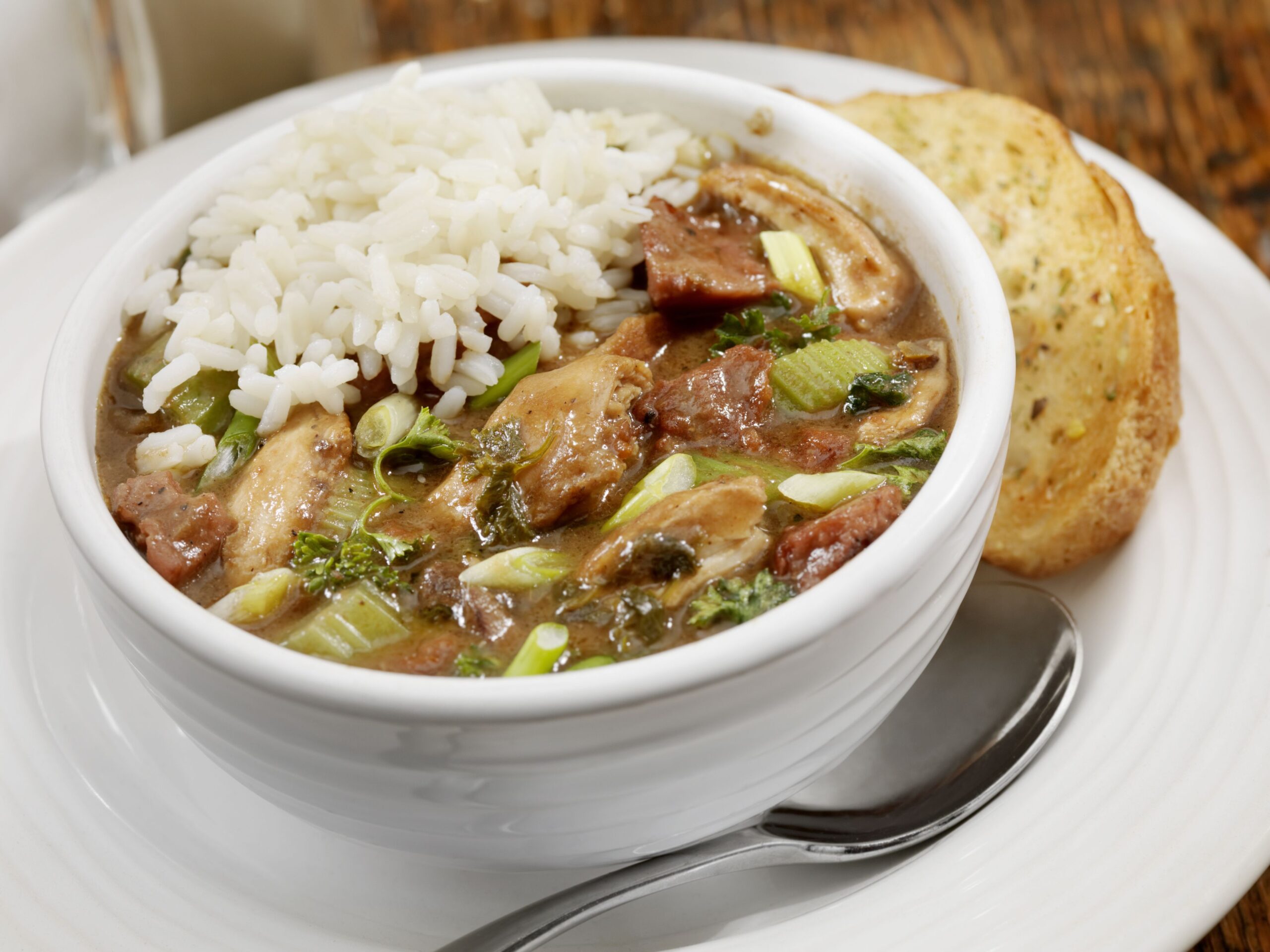Closeup of chicken gumbo with rice in a white bowl with a spoon and a slice of garlic bread on a matching white saucer, with a blurred background of a rustic wooden table