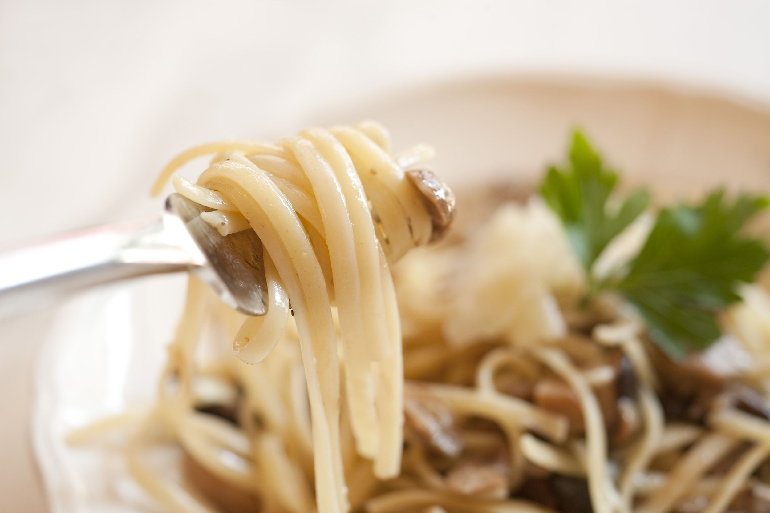 Closeup of linguine with mushrooms and parmesan cream sauce on a fork with fancy porcelain plate of it blurred in the background, against a white tablecloth