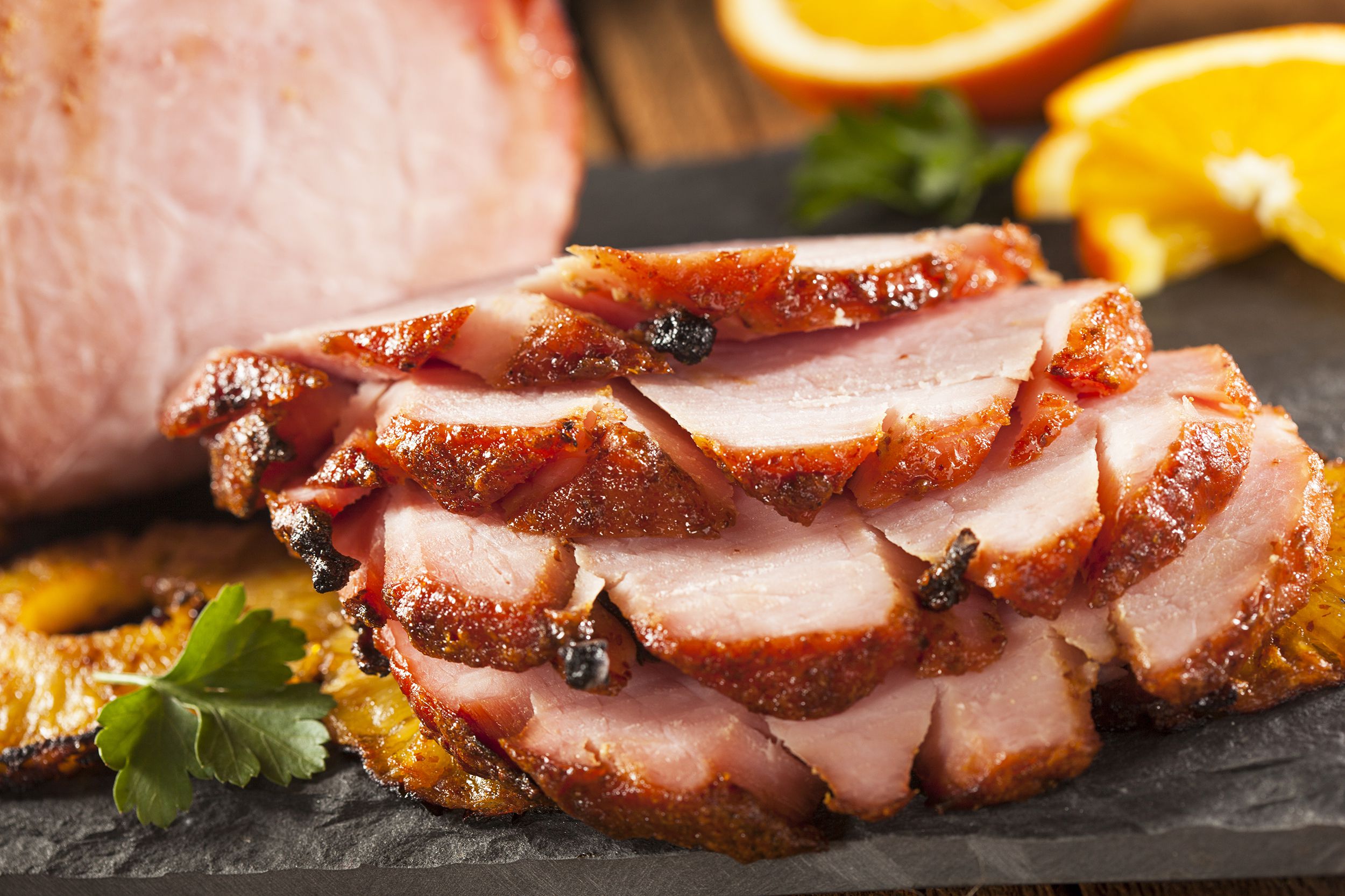 Closeup of five thick slices of simple baked ham in the foreground on a slate serving board with rest of the ham and slices of orange, blurred in the background