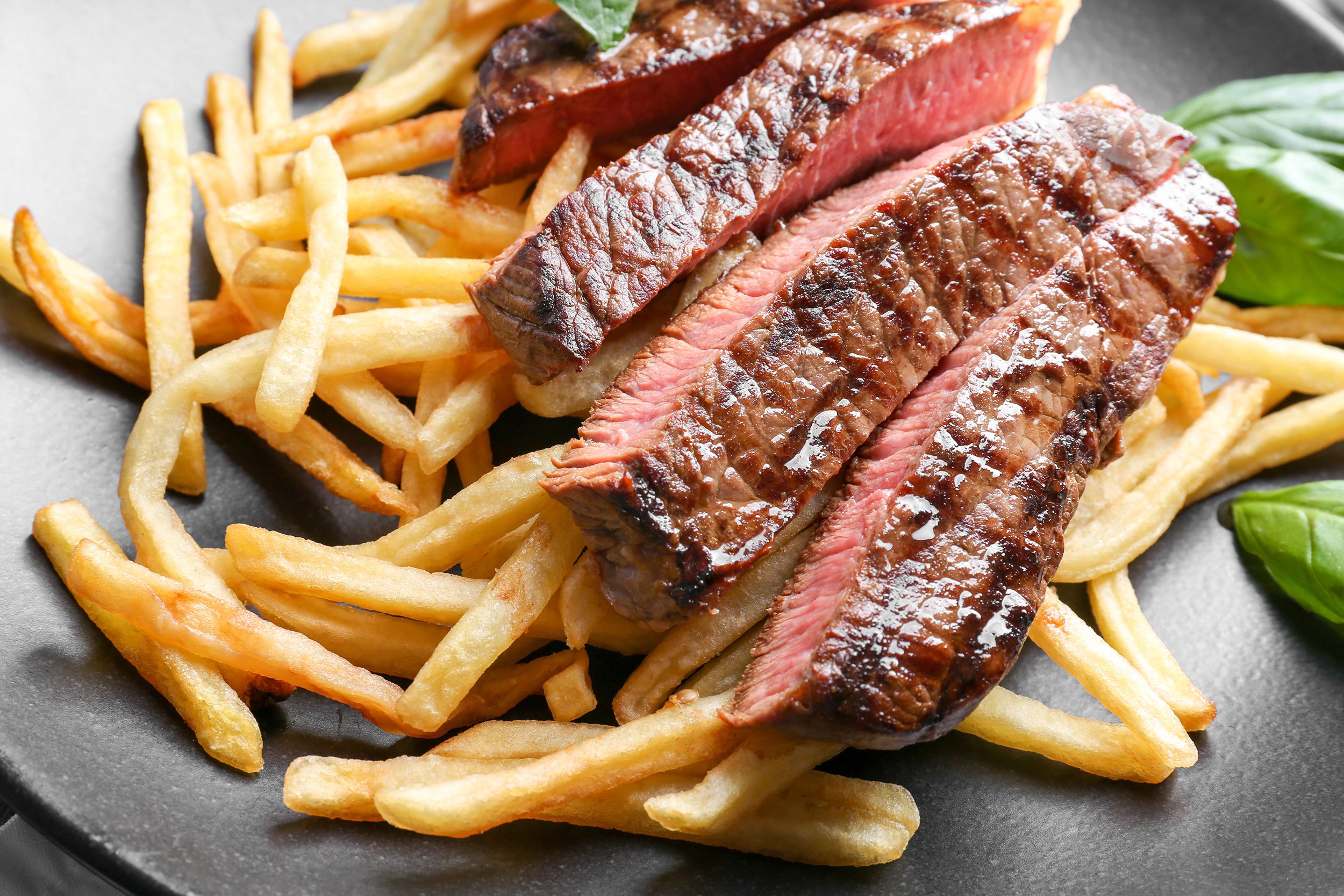 Closeup of four slices of steak frites on slim French fries on a black ceramic plate with a few herb leaves on the right