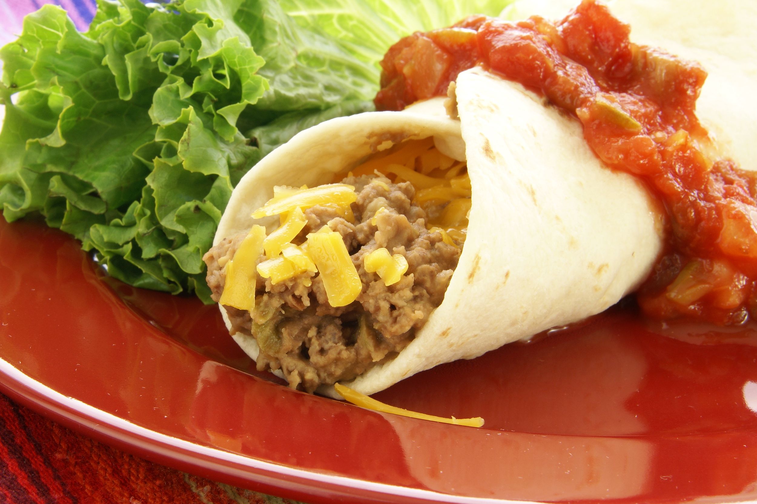 Closeup of one refried bean burrito with sauce on top, next to lots of lettuce, on a red plate with a red tablecloth in the left corner
