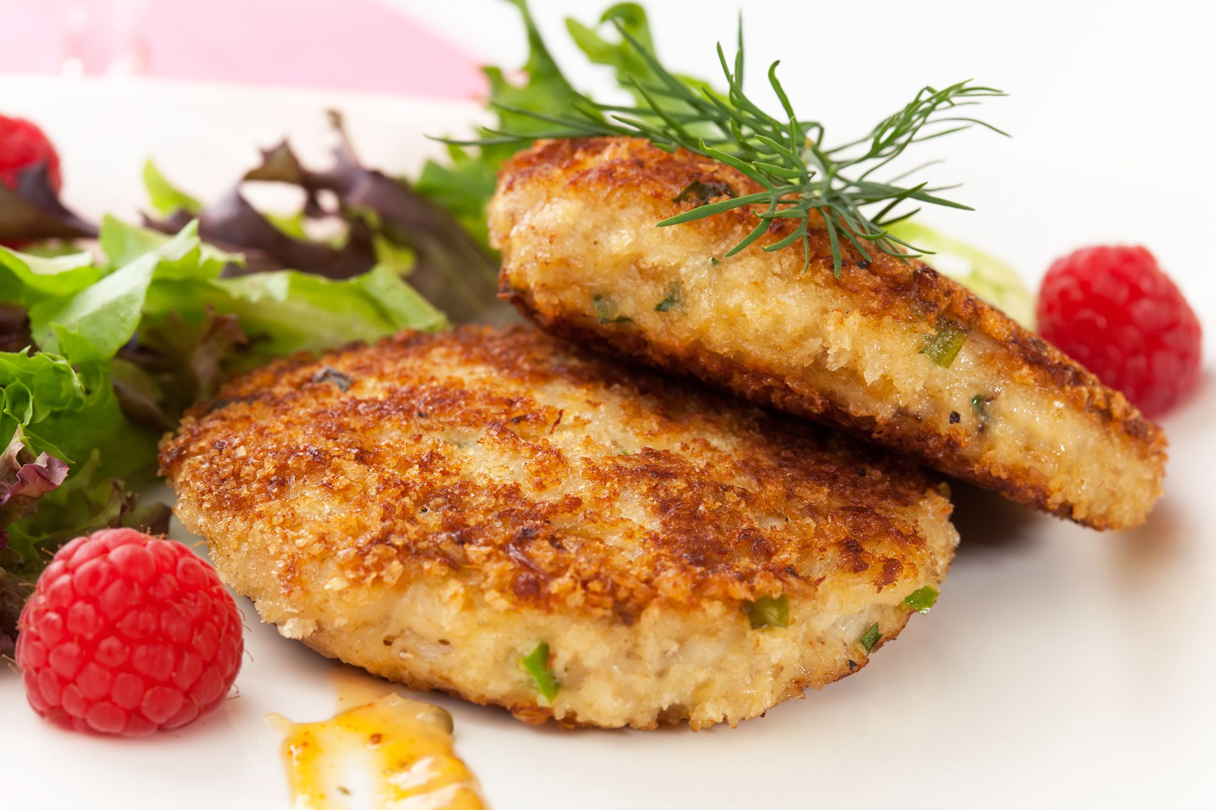 Closeup of two Maryland crab cakes stacked on one another, surrounded by arugula and raspberries, on a white plate with a slight blurred background