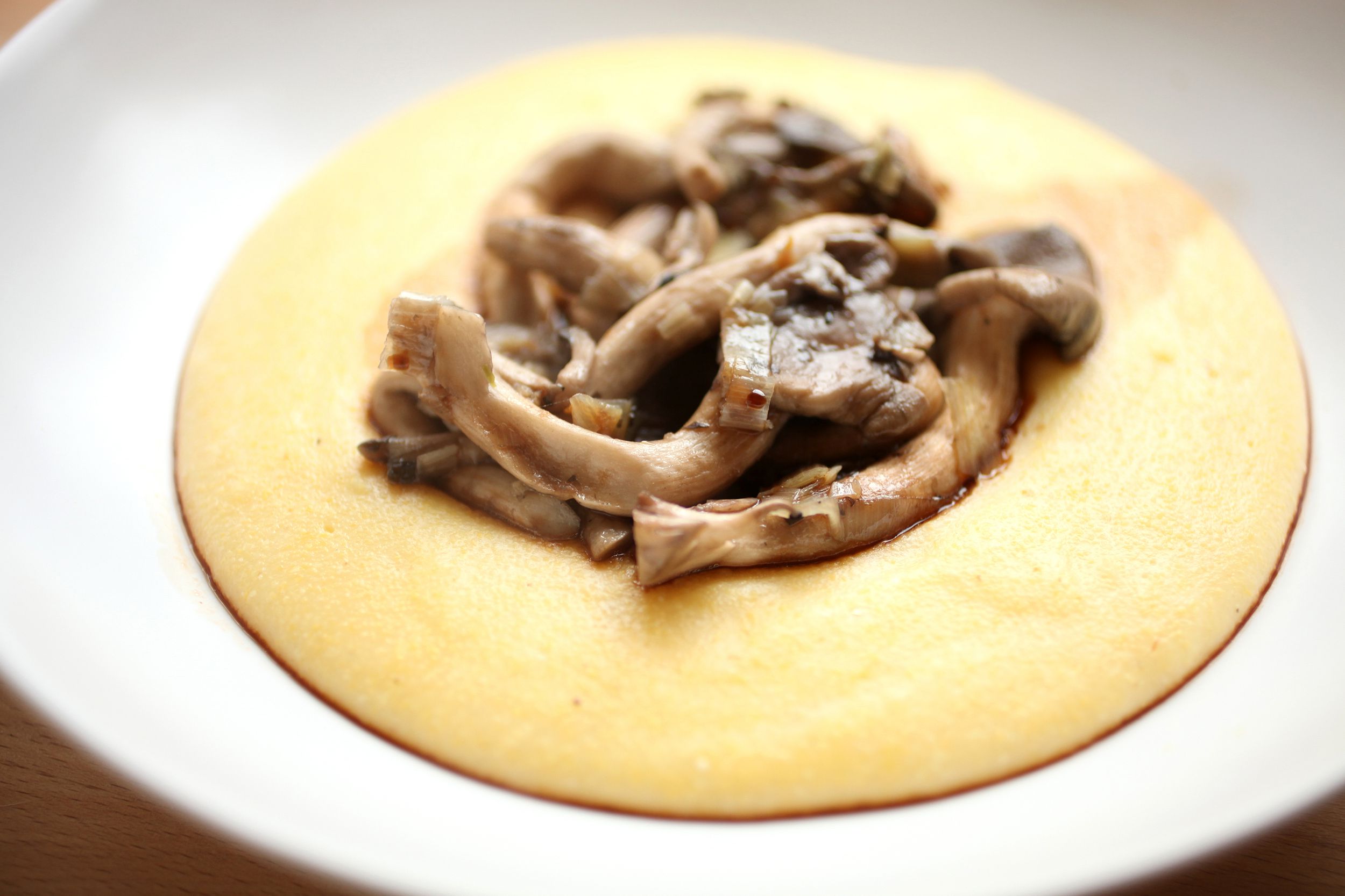 Closeup of wild mushroom ragout over creamy polenta in a white porcelain bowl with a blurred background of a table