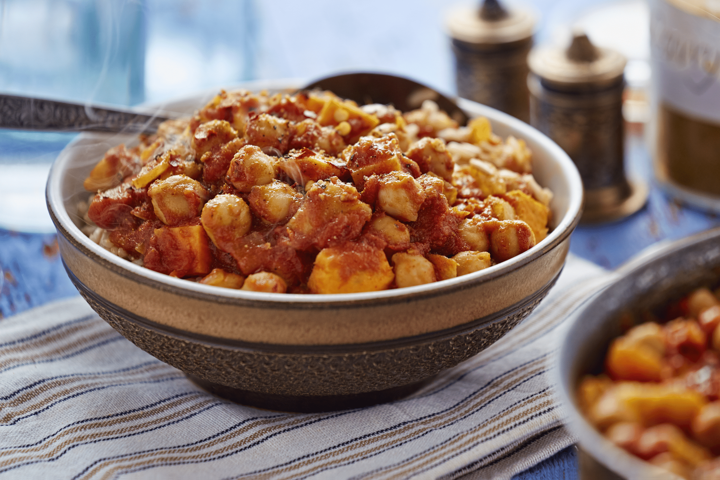 Creamy vegan curry in a lined brown ceramic bowl with stem coming off of it, a spoon in it, selective focus, on a brown and beige lined napkin, surrounded by another bowl, salt and pepper shakers, and more items blurred in the background