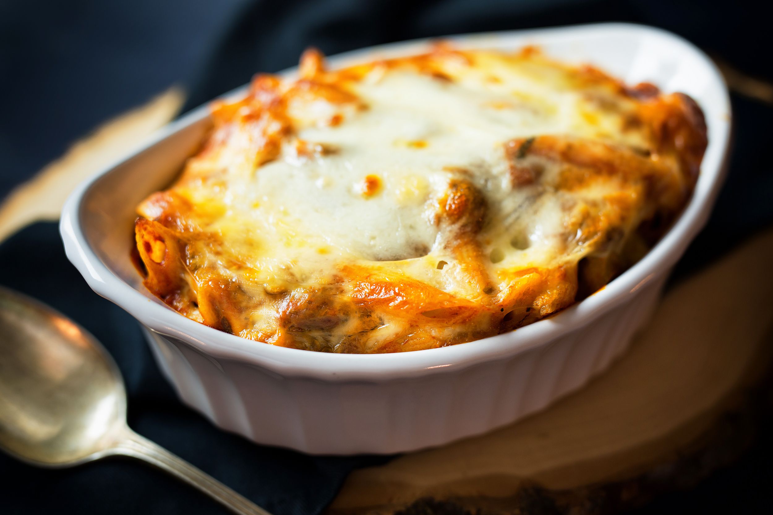 Easy baked ziti in a white porcelain dish, selective focus, a spoon to the left with a blurred background of a serving board and a napkin with shading