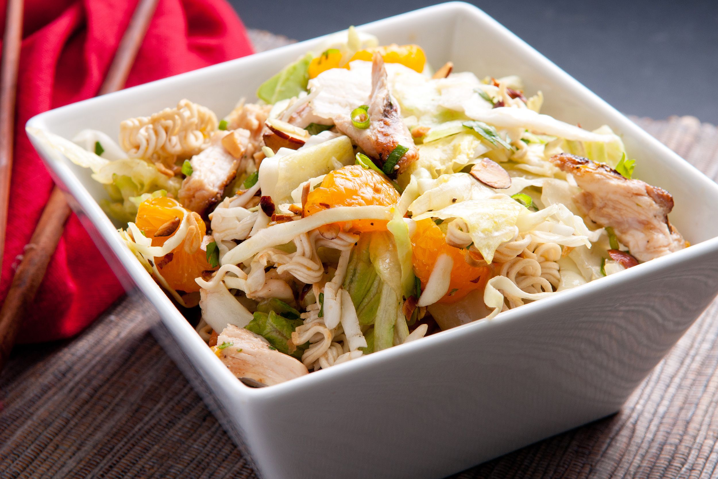 Easy spicy Asian chicken salad in a square white bowl with a red napkin and two wooden chopsticks on a table mat