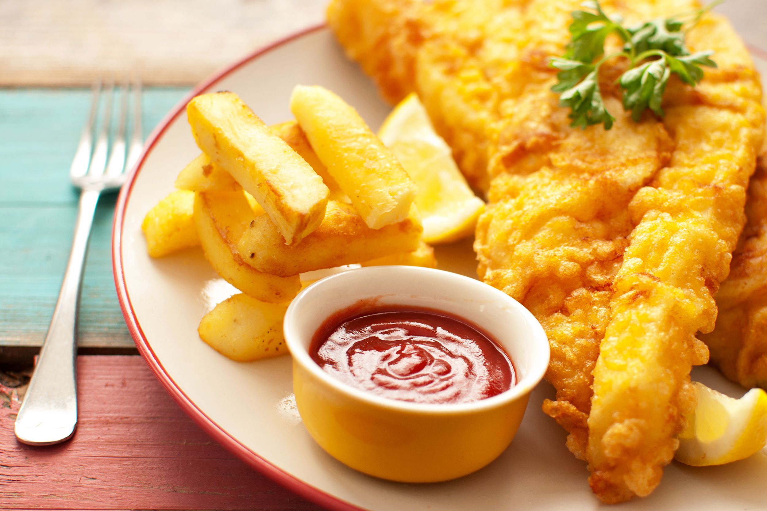 Fish and chips on a red rimmed white plate with ketchup in a small yellow and white inside dipping bowl with a fork on the left next to the plate, on a blue and red wooden table with a blurred background