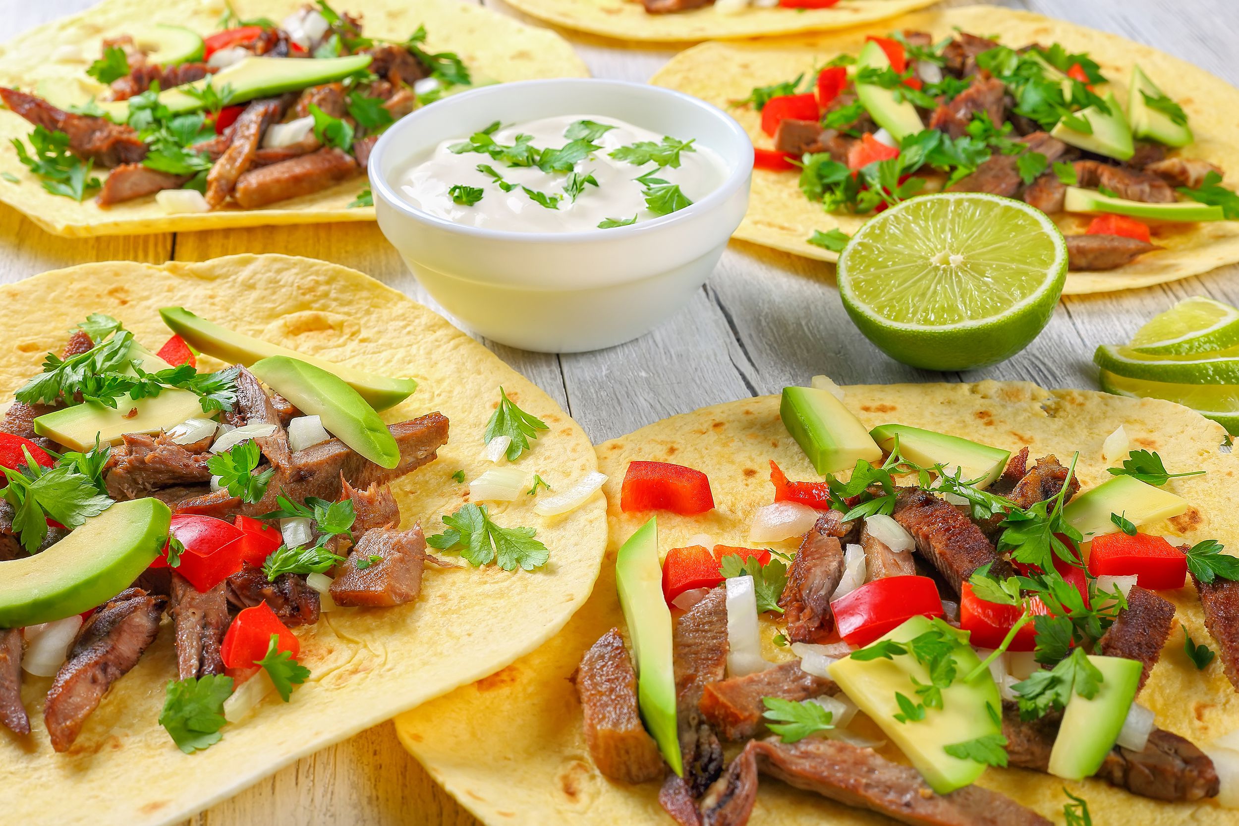 Five delicious beef tongue tacos in a circle around a small bowl of white dipping sauce, slight horizontal view, on a white on a table