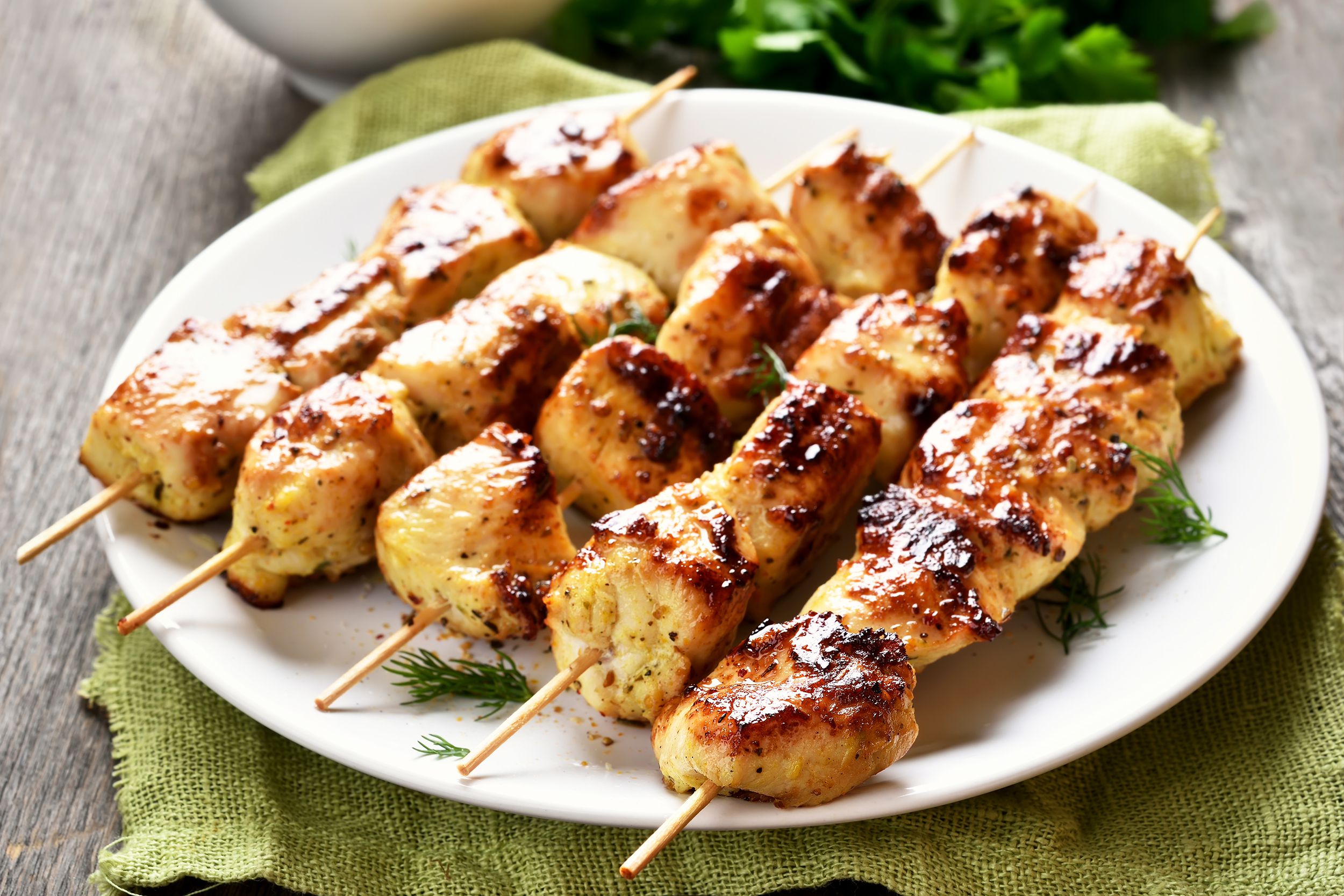 Five middle eastern-style grilled chicken kabobs on a white plate, selective focus, on a avocado green napkin, next to lettuce, on a grey wooden table