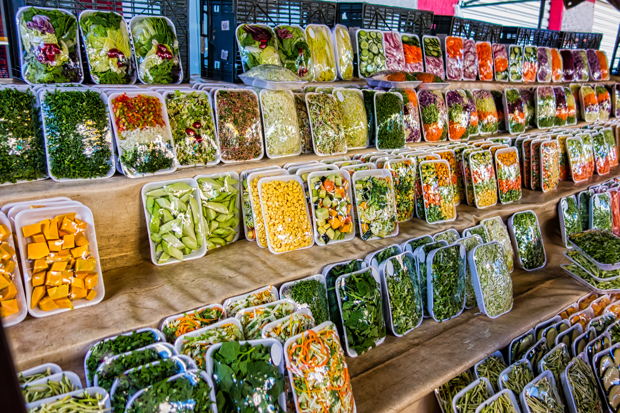 Five rows of packaged vegetables, different kinds, mixes, salads, all in white Styrofoam and plastic wrapped, on wooden shelves in a semi-pyramid