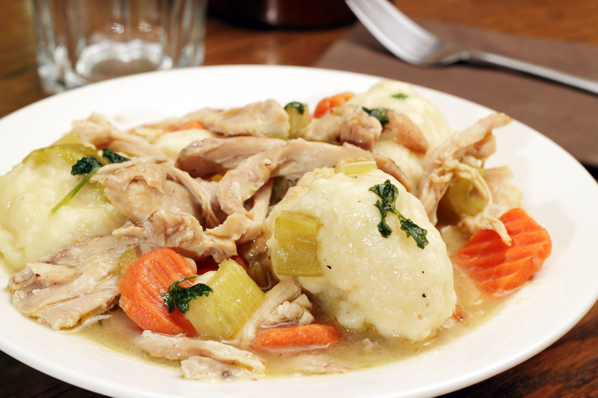 Focus on classic chicken and dumplings in a white bowl with a fork and a glass of water on a table in the background