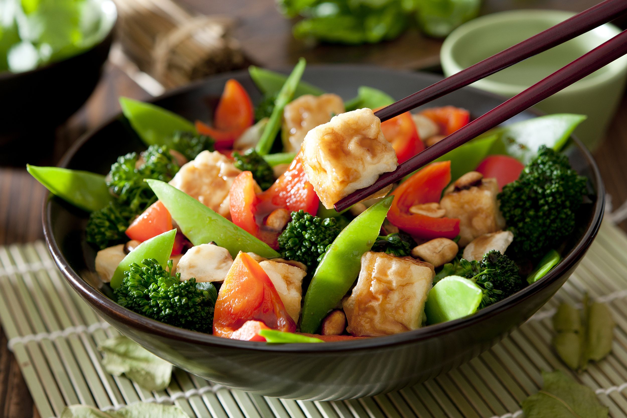 Honey ginger tofu and veggie stir fry in a black Asian bowl with black chopsticks holding a piece of tofu over it on a bamboo sushi mat, with another bowl and ingredients on a wooden table, blurred in the background