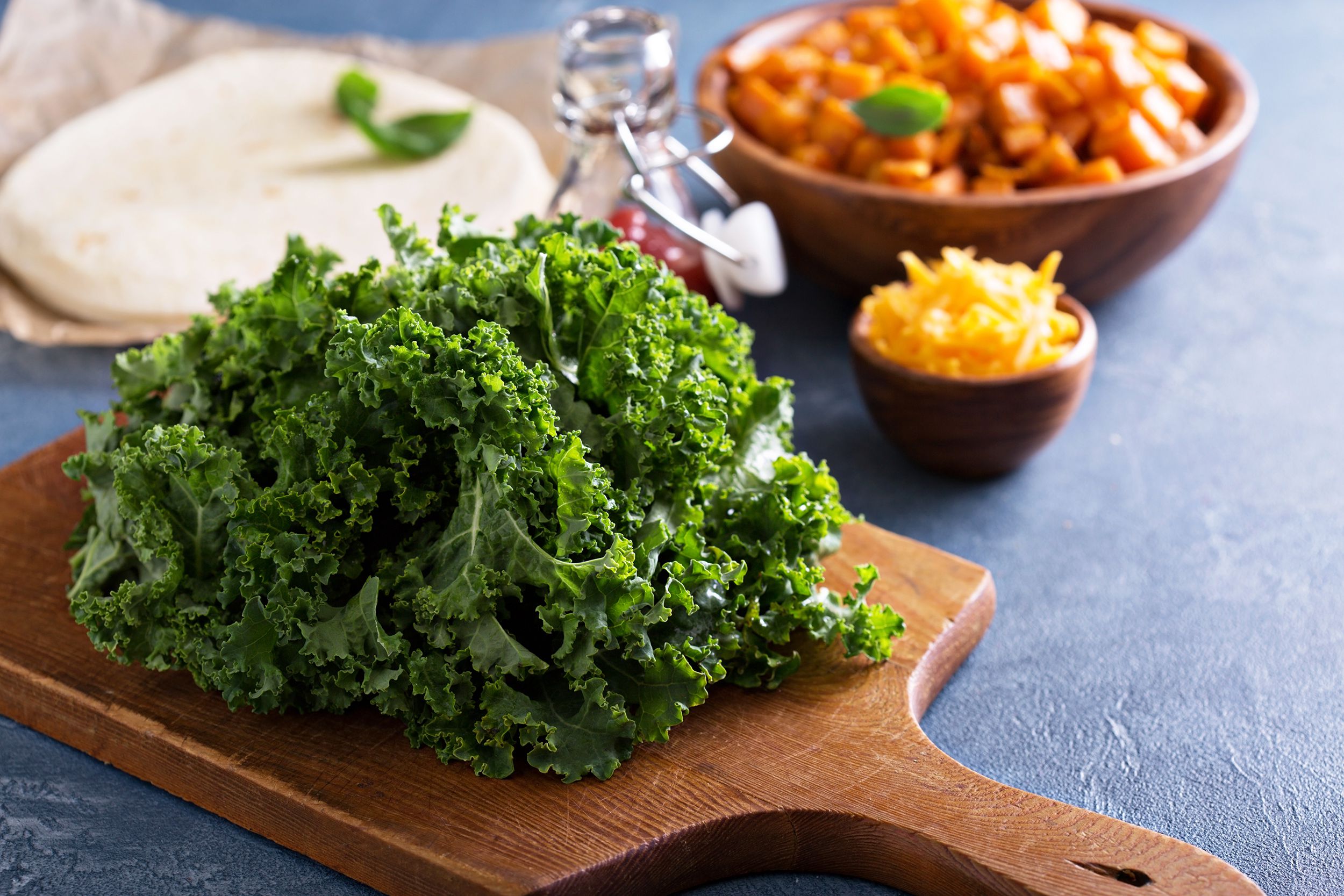 Ingredients of sweet potato tacos without beans, a wooden cutting board of lettuce next to soft taco shells, a ceramic bowl of sweet potatoes, a small ceramic bowl of cheese, and a glass bottle of sauce on a blue table