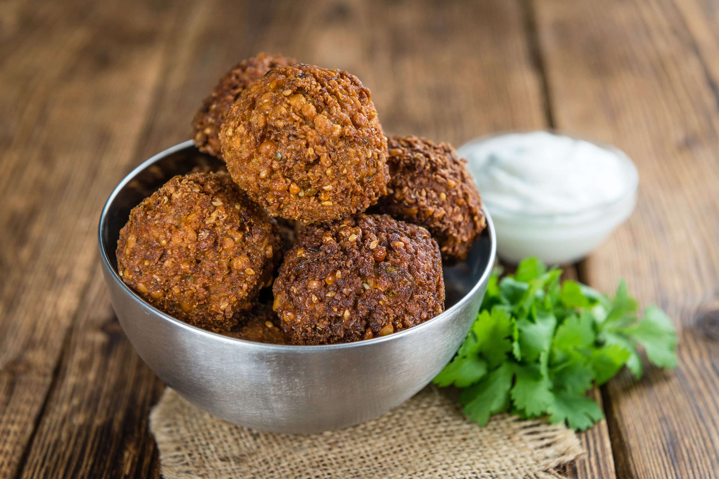 Many falafel piled in a stainless steel bowl, selective focus, on a natural napkin with herbs and white dipping sauce with a blurred background of a rustic wooden table