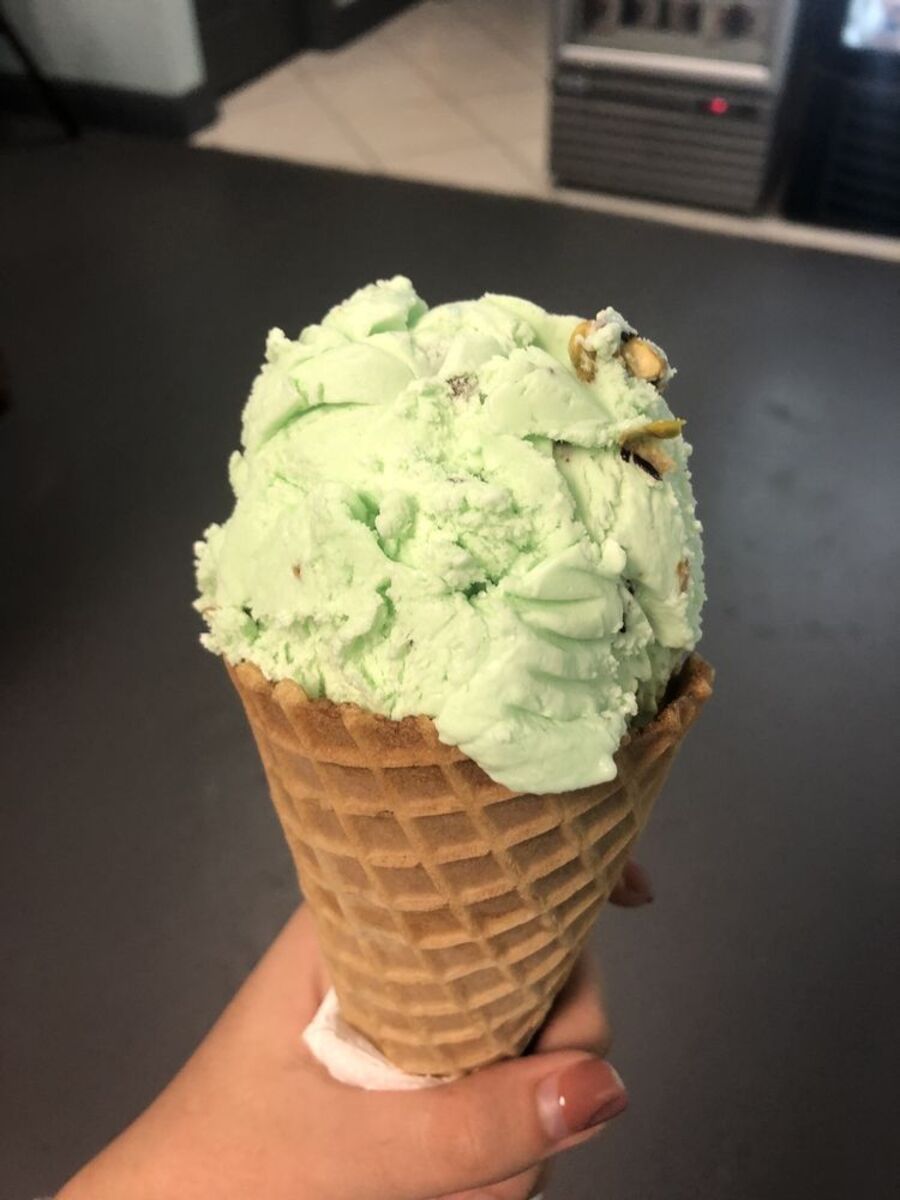 Pistachio ice cream on a waffle cone, Big Olaf Creamery, Sarasota, Florida, hand holding it, grey countertop in the background with creamery machines and beige floor at the top