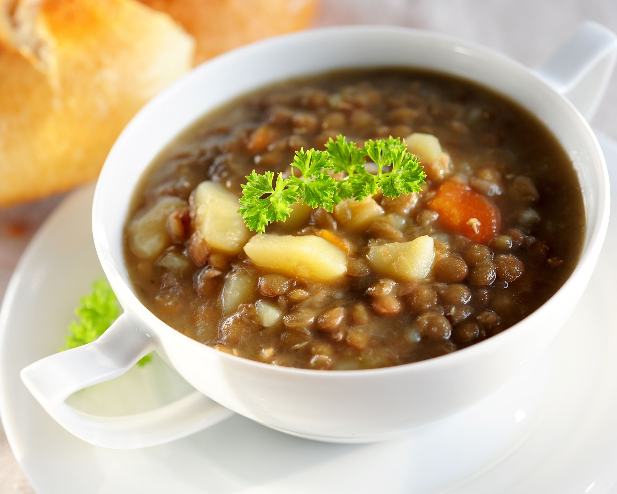 Pressure cooker lentil soup in a porcelain white bowl with two handles on a matching porcelain white plate, selective focus, with two small loaves of bread