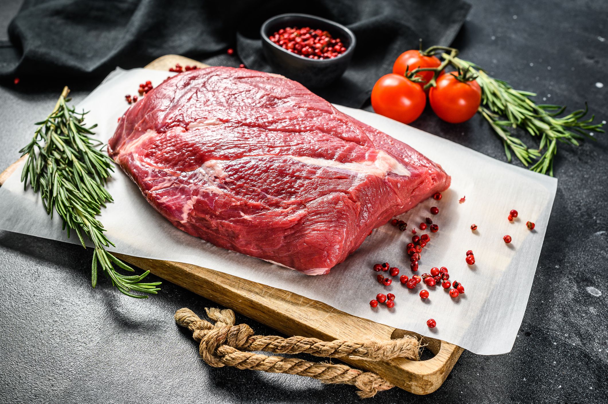Raw brisket angus beef slab on a sheet of wax paper on a wooden cutting board on a slate grey table, surrounded by tomatoes and herbs
