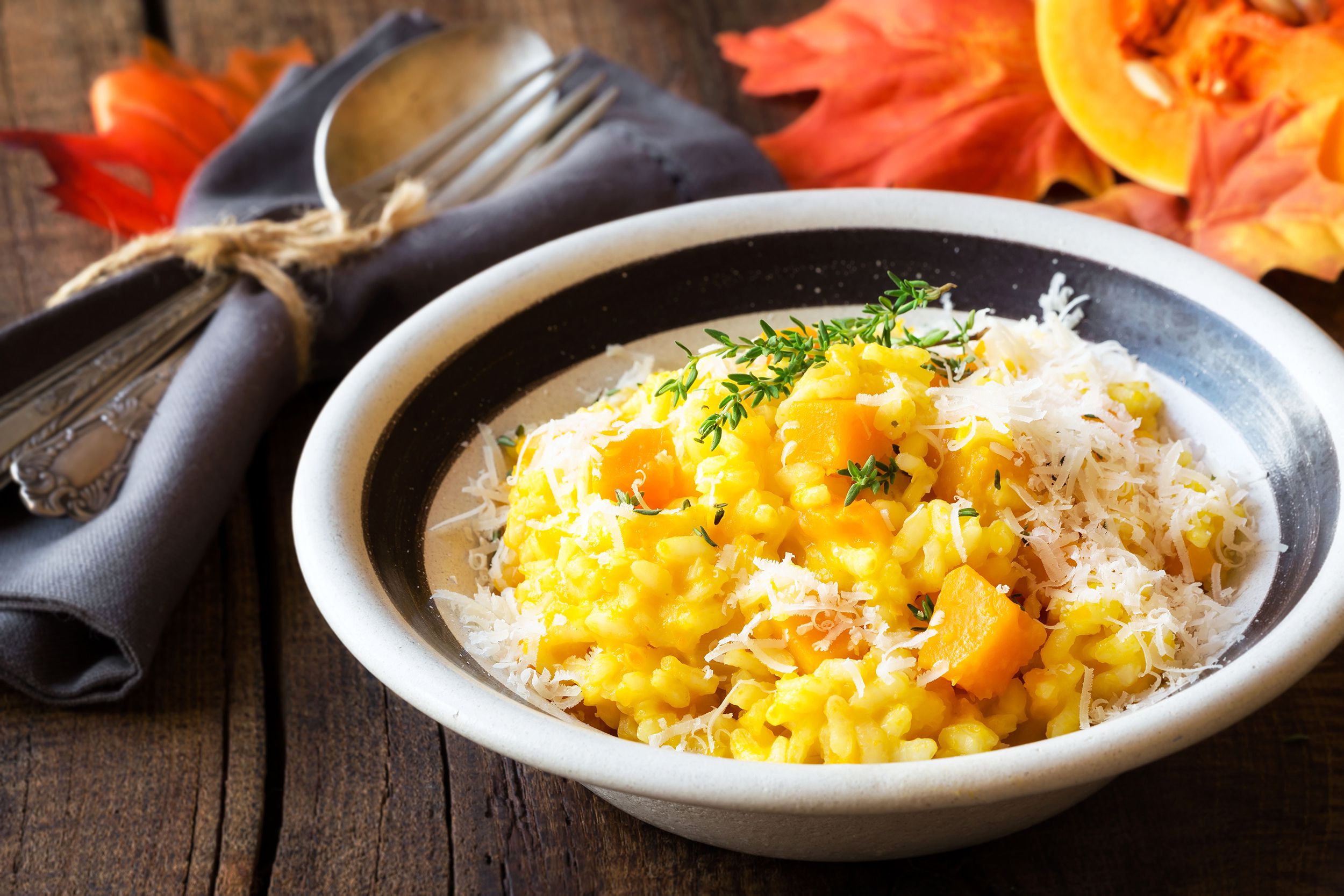 Roasted butternut squash risotto in a black lined white ceramic bowl, selective focus, with a fork and spoon tied in a navy napkin to the left on a rustic wooden table with autumn leaves in the background