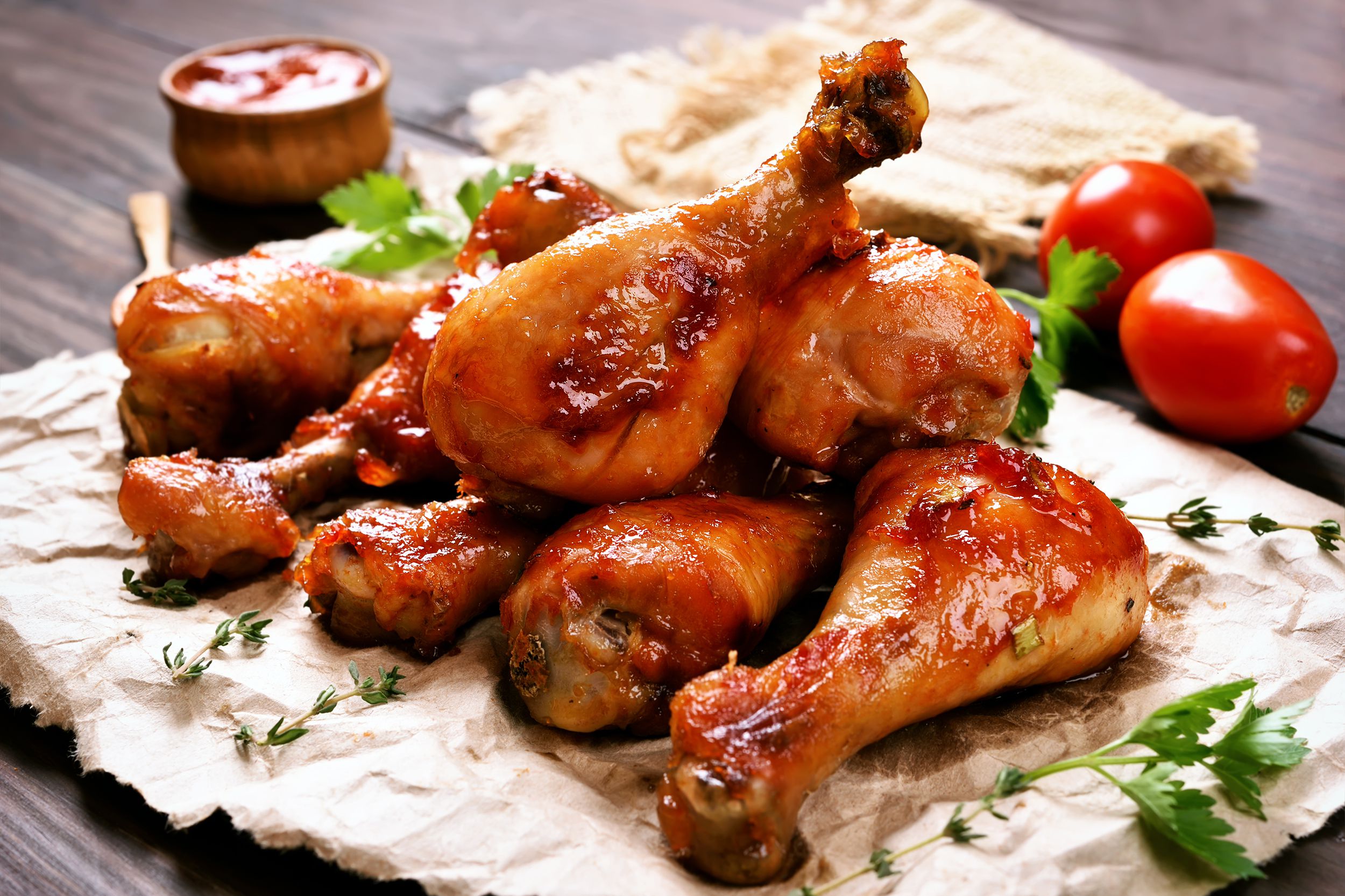 Seven sweet & spicy roasted chicken legs piled together on rustic brown paper on a dark brown wooden table with sauce in a bowl and a spoon, blurred in the background
