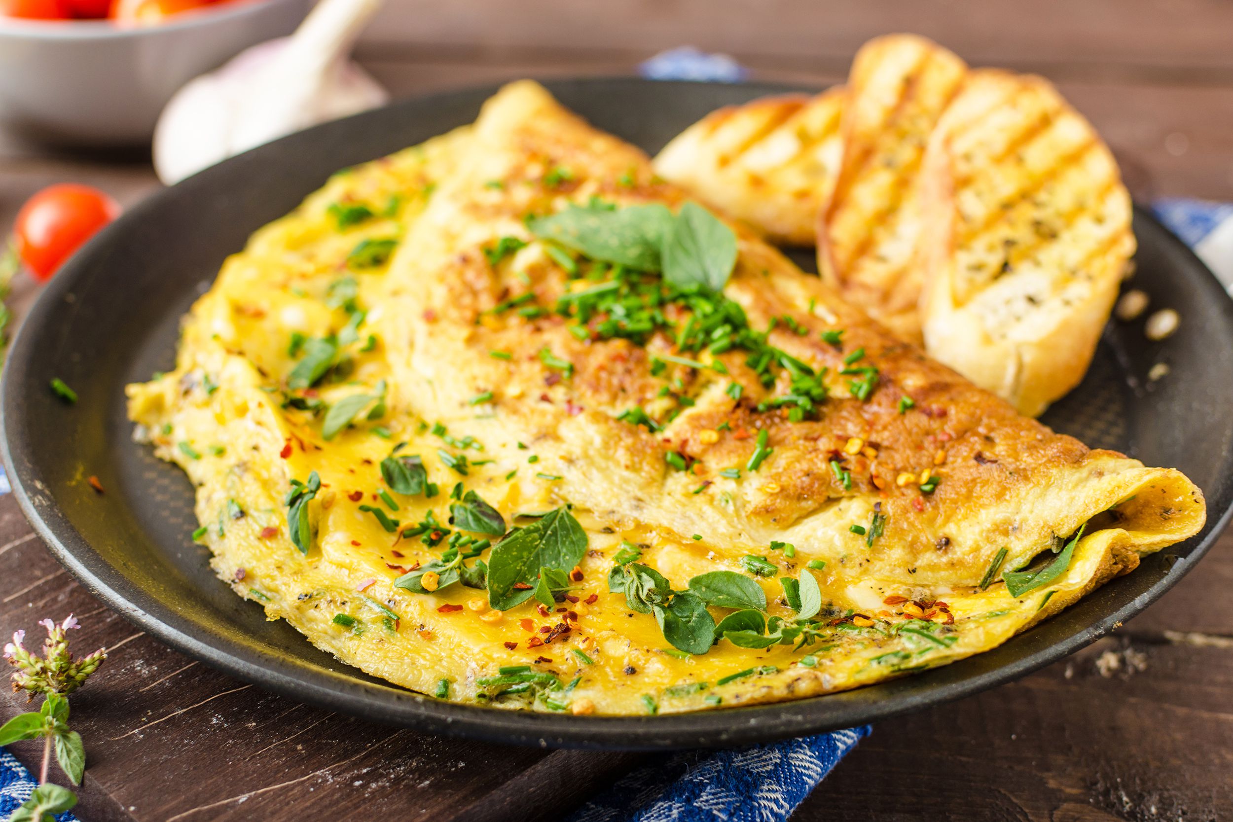 Simple cheese omelette in a black ceramic plate, selective focus, with three pieces of garlic bread on a dark brown wooden table, with tomatoes and garlic in the background