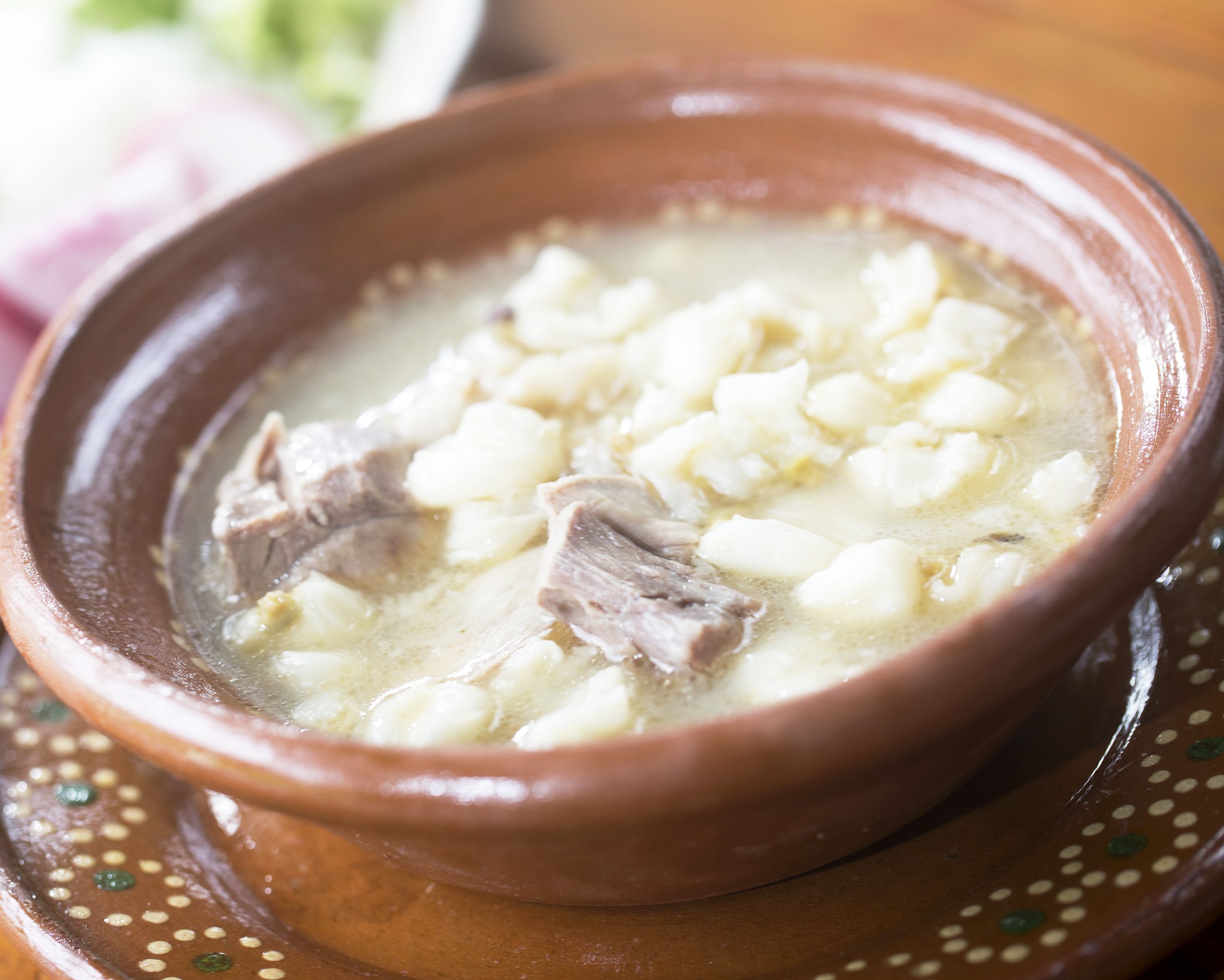 Simple pork posole in a terracotta bowl on a matching decorative saucer, selective focus, with a blurred background of ingredients and table