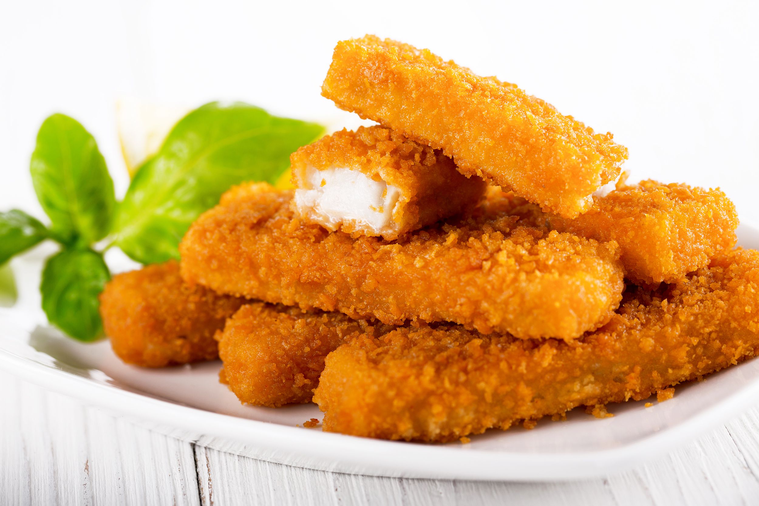 Six homemade fish sticks stacked on a white porcelain plate on a white table, with a blurred background