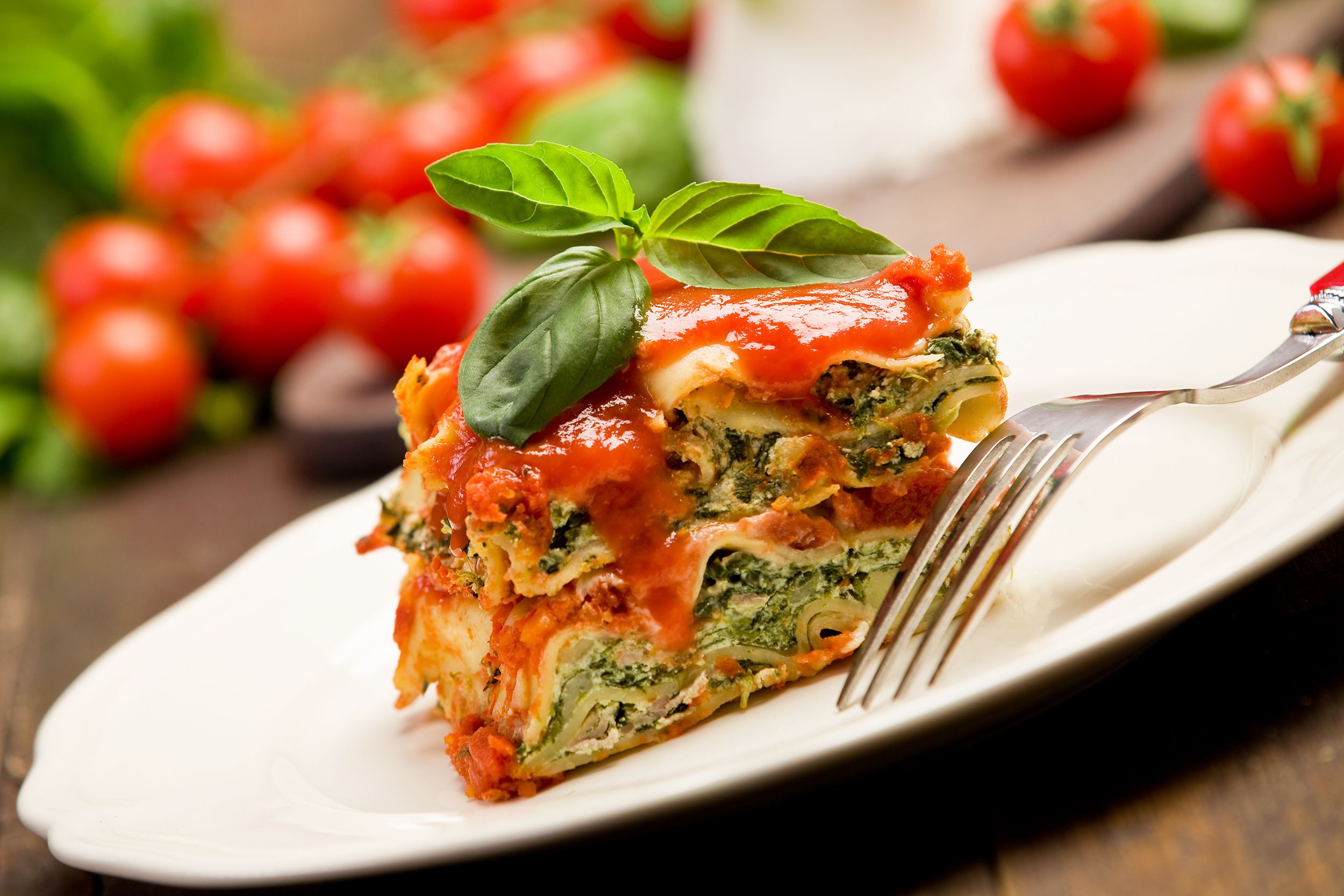A slice no-fuss vegetarian spinach lasagna on a white porcelain plate with a fork, diagonal, selective focus, with tomatoes and ingredients blurred in the background